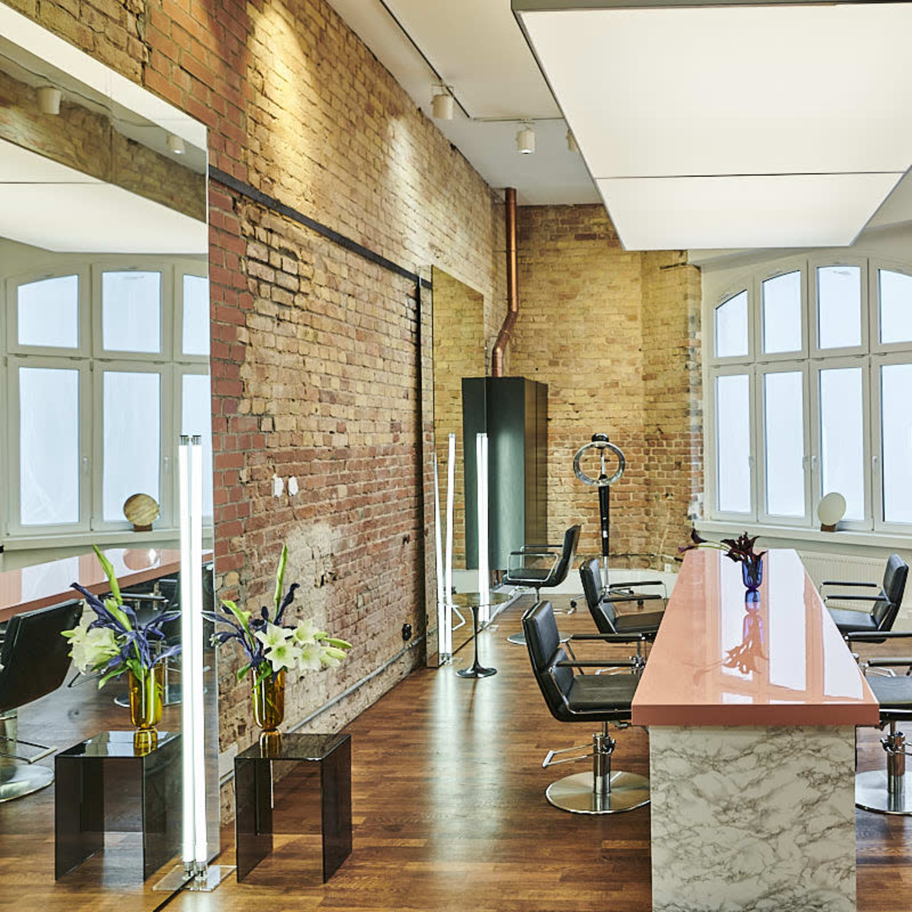 A modern salon interior with a pink marble table, black chairs, and large windows along a brick wall.