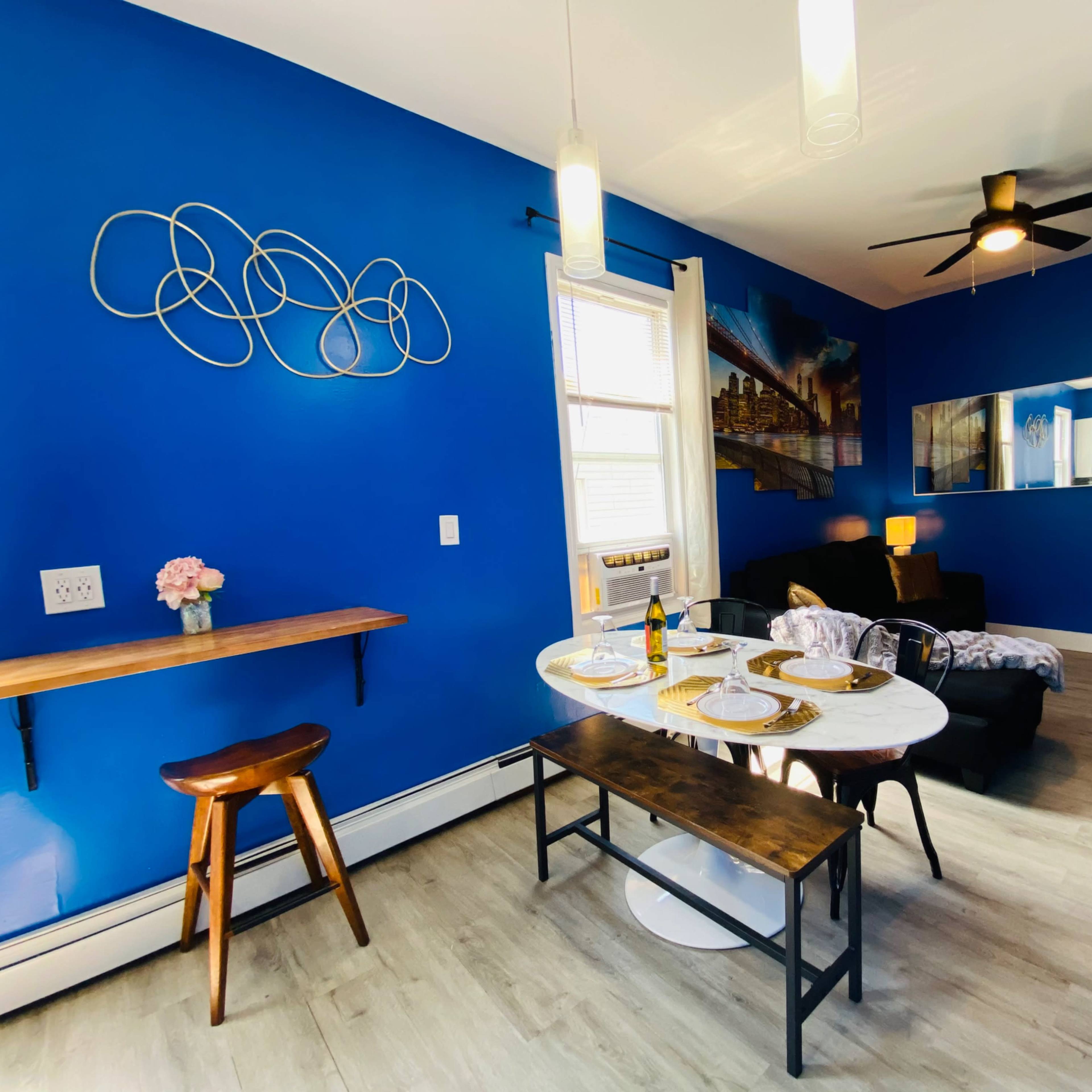 A dining area with a blue wall, a small table set for a meal, a wooden bench, and a living room visible in the background.