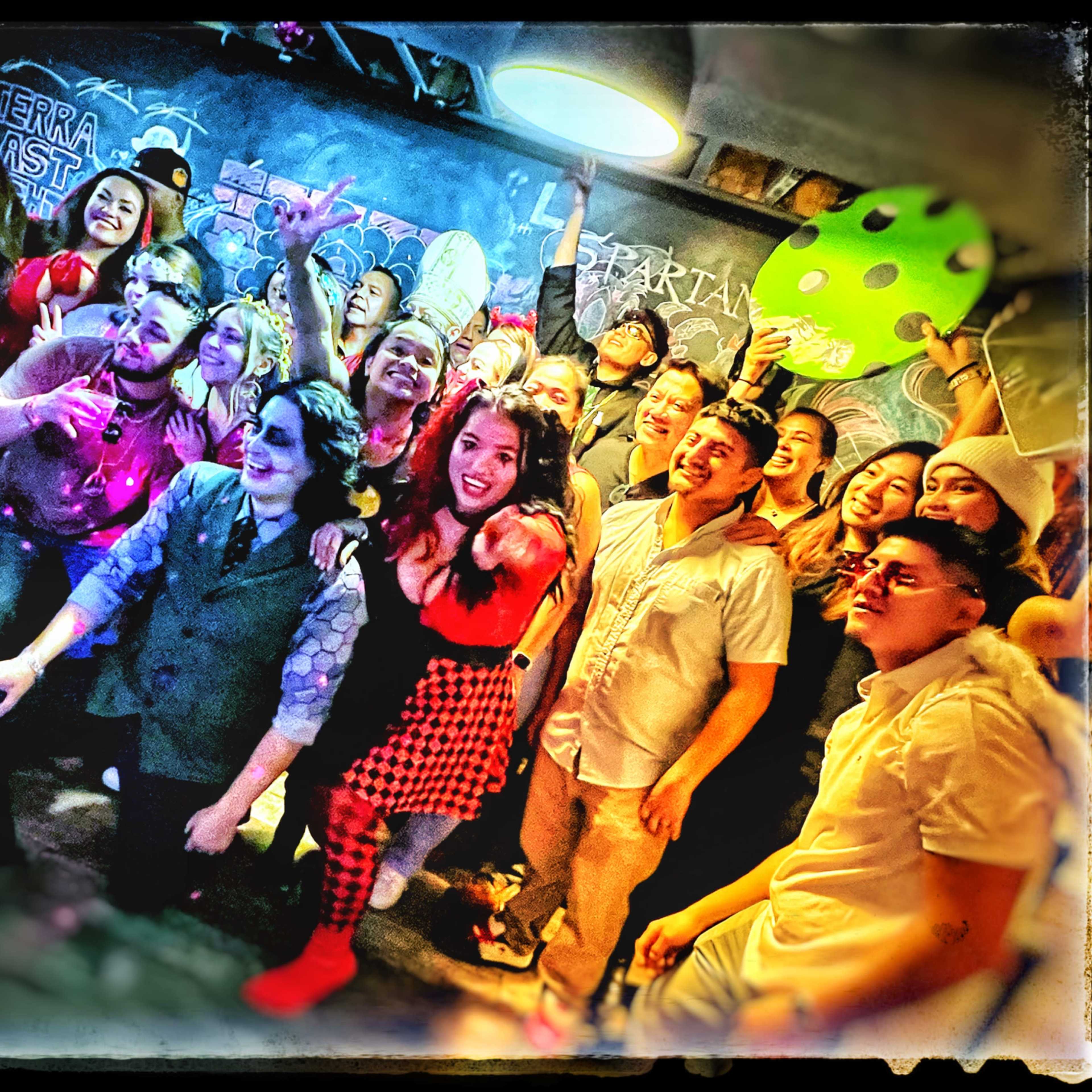 A diverse group of people poses together with cheerful expressions in a dimly lit venue, some holding props and costumes.