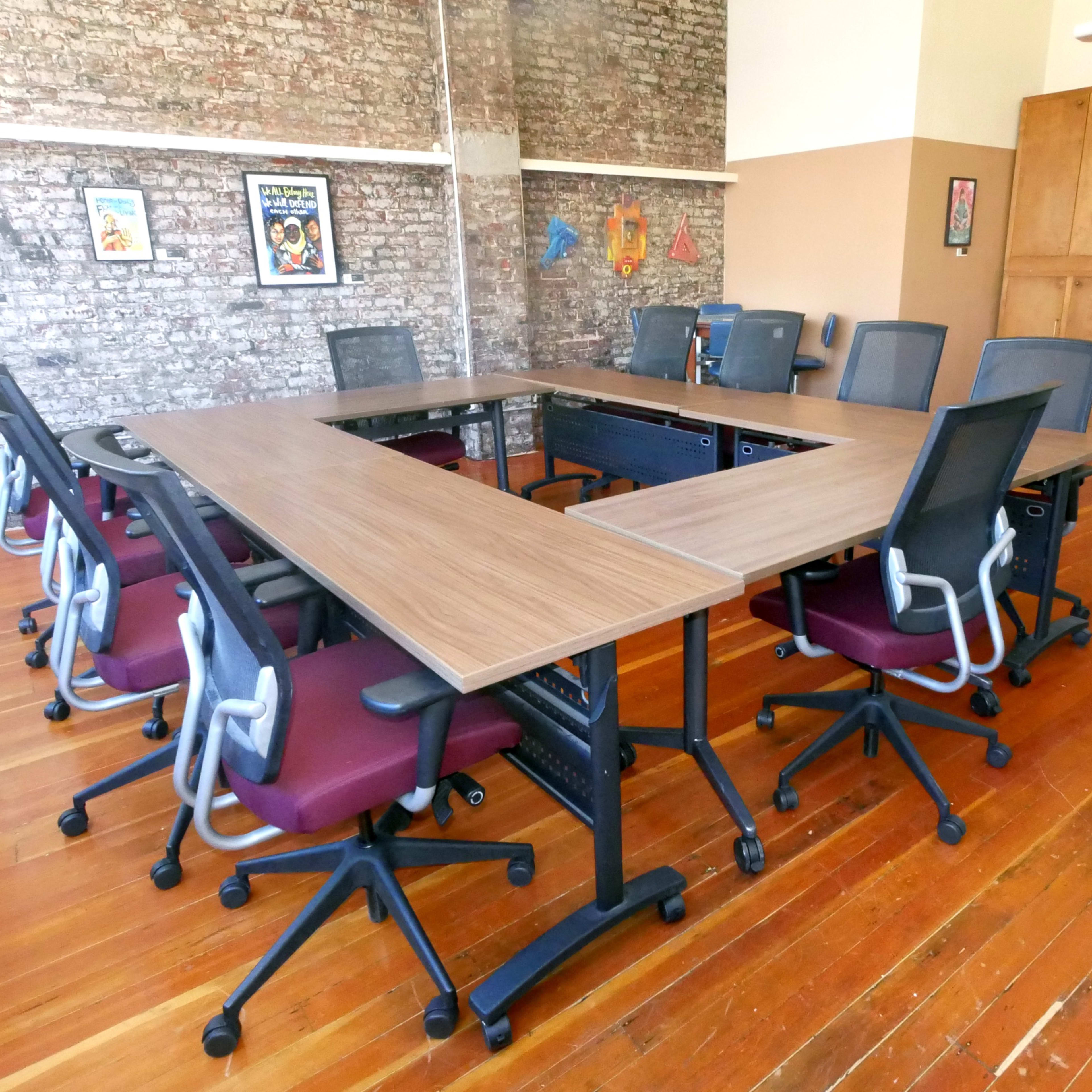 A conference room features a large rectangular table surrounded by several chairs, against a backdrop of brick walls adorned with posters.