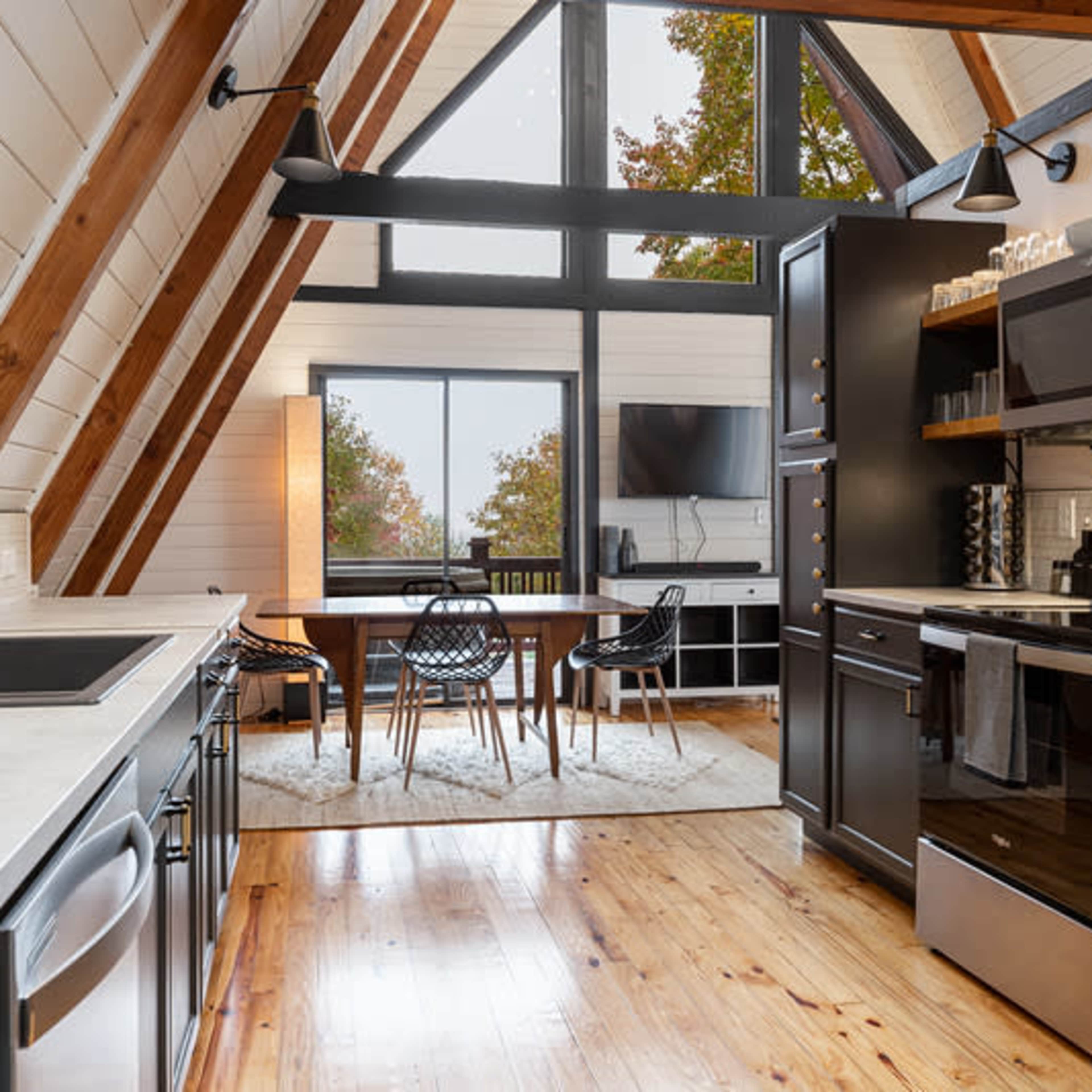 A modern kitchen with a high vaulted ceiling, featuring wooden beams, a large window, and a dining area with a black table and chairs.