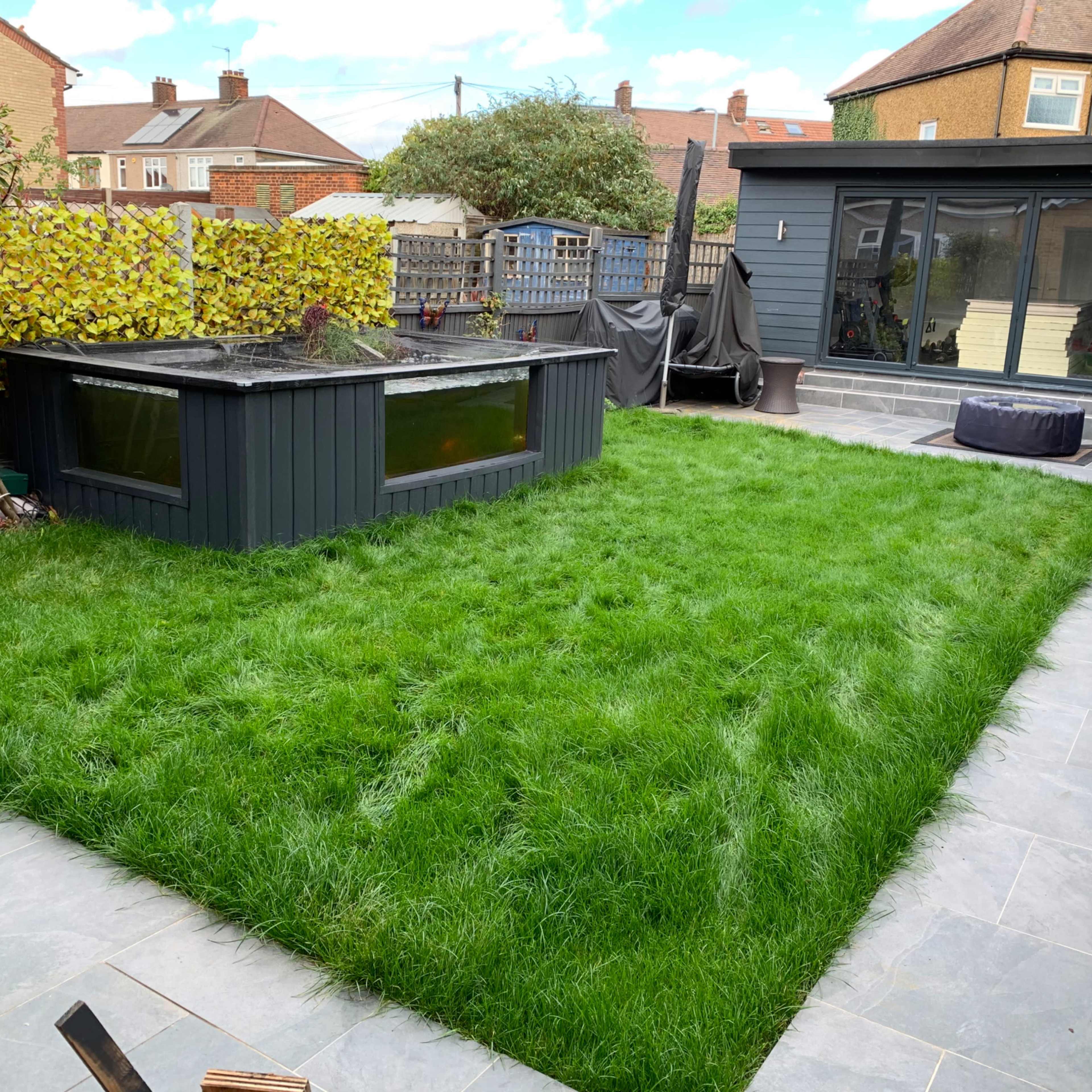 A landscaped yard features a green lawn, a dark-colored garden shed with a water feature, and a patio area with slate tiles.