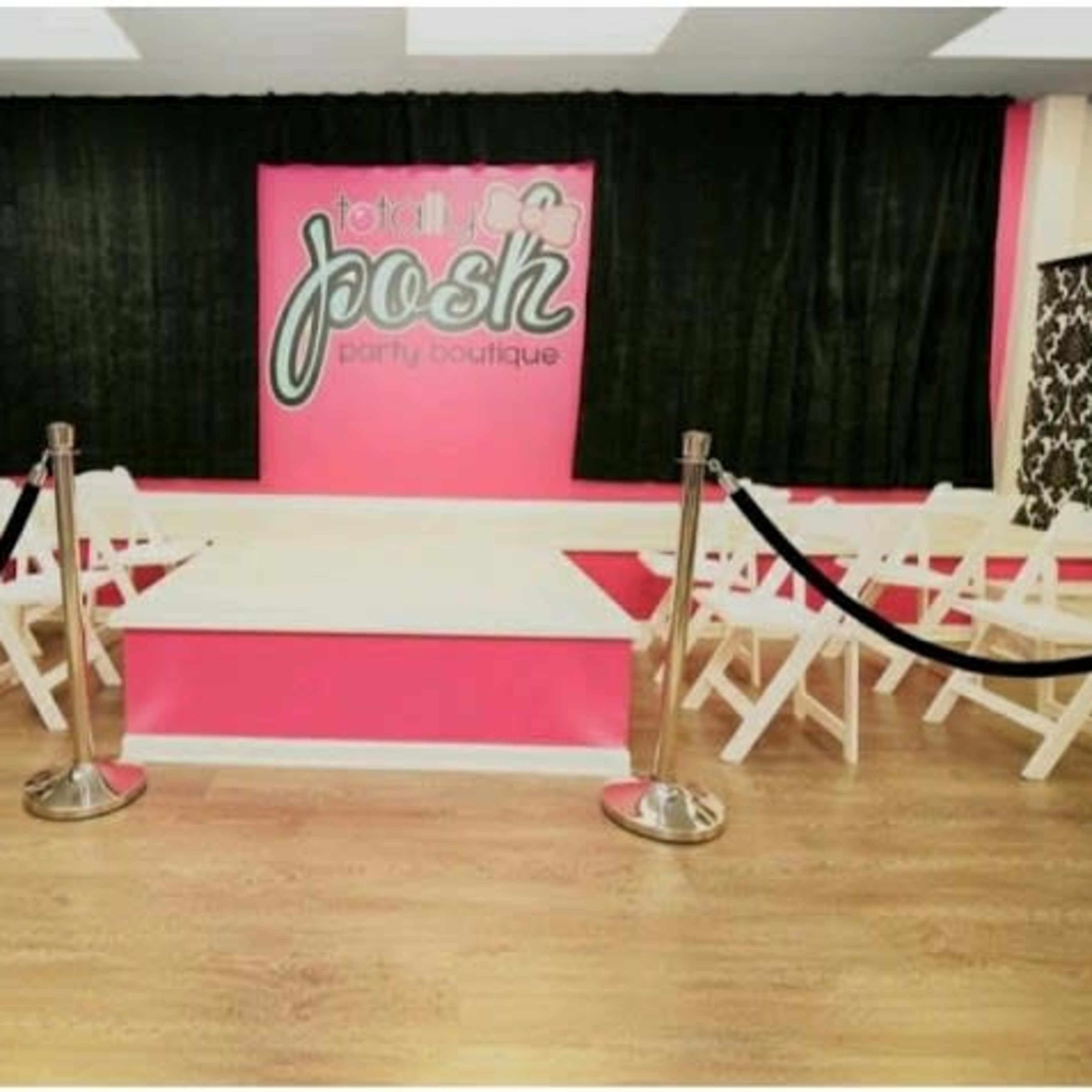 A party boutique interior featuring a runway-style stage, white folding chairs arranged in front, and a vibrant pink and black color scheme on the walls.