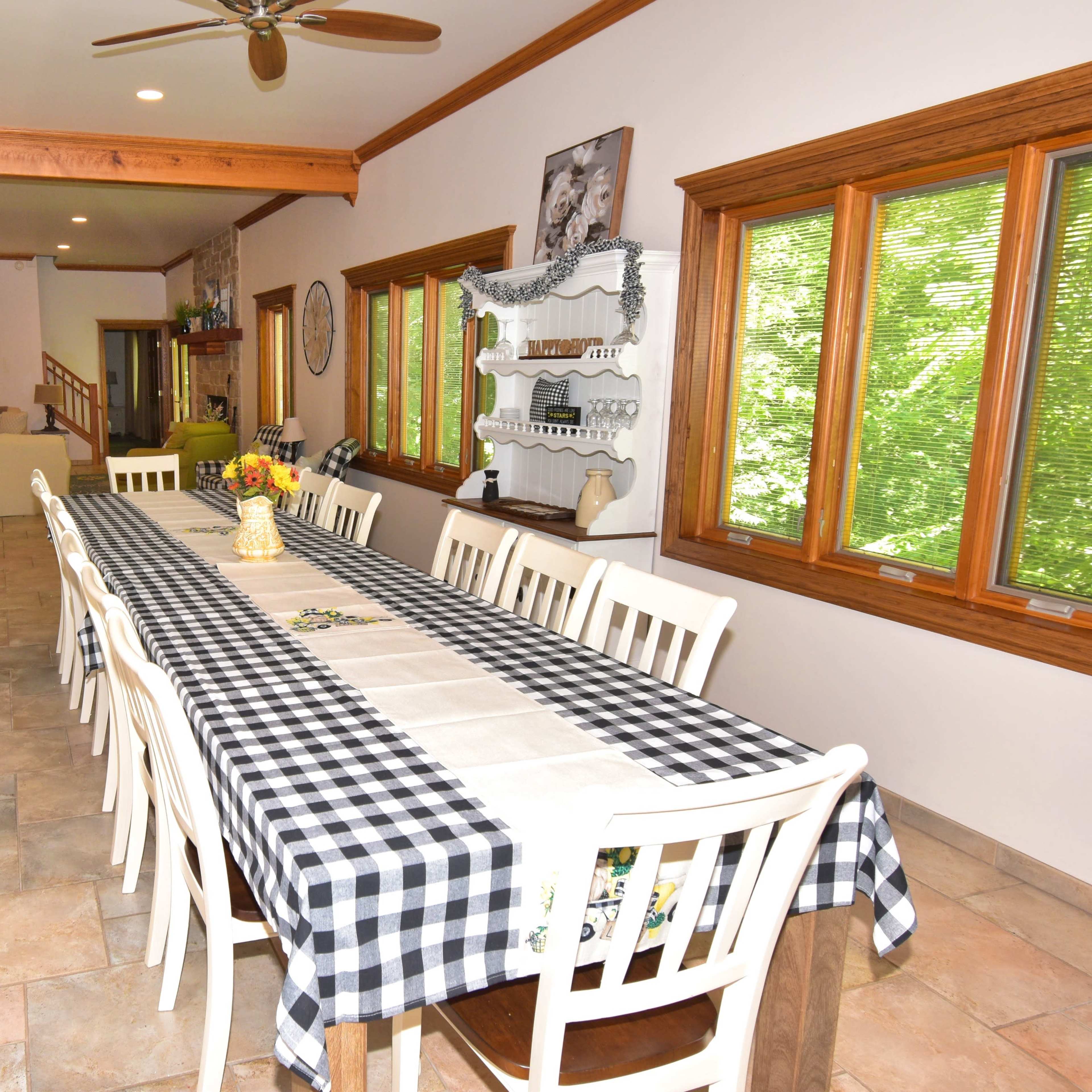 A long dining table with checkered tablecloth is set in a spacious room with large windows and a view of greenery outside.
