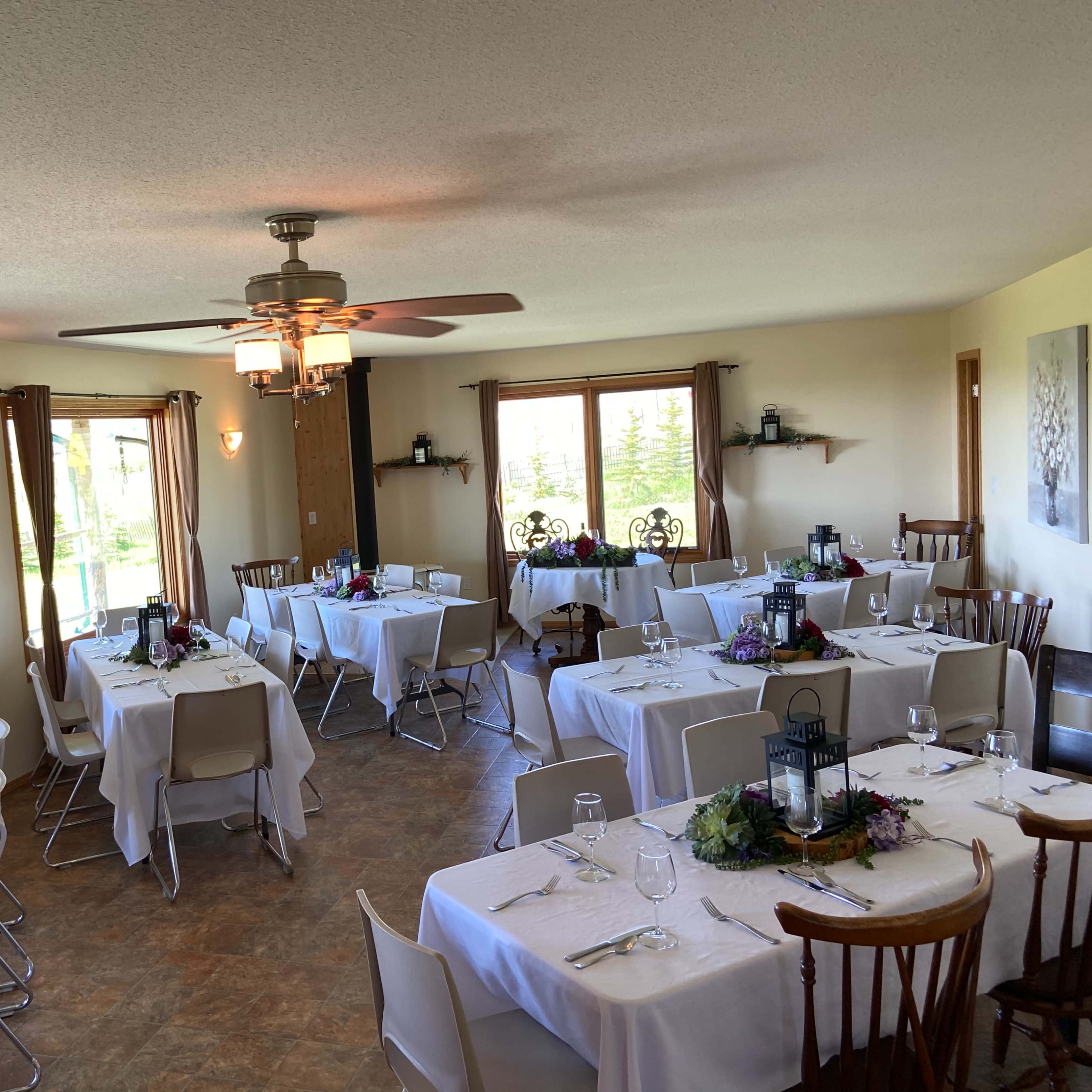 A dining area with several tables set for a meal, featuring white tablecloths, utensils, and centerpieces, under a ceiling fan.