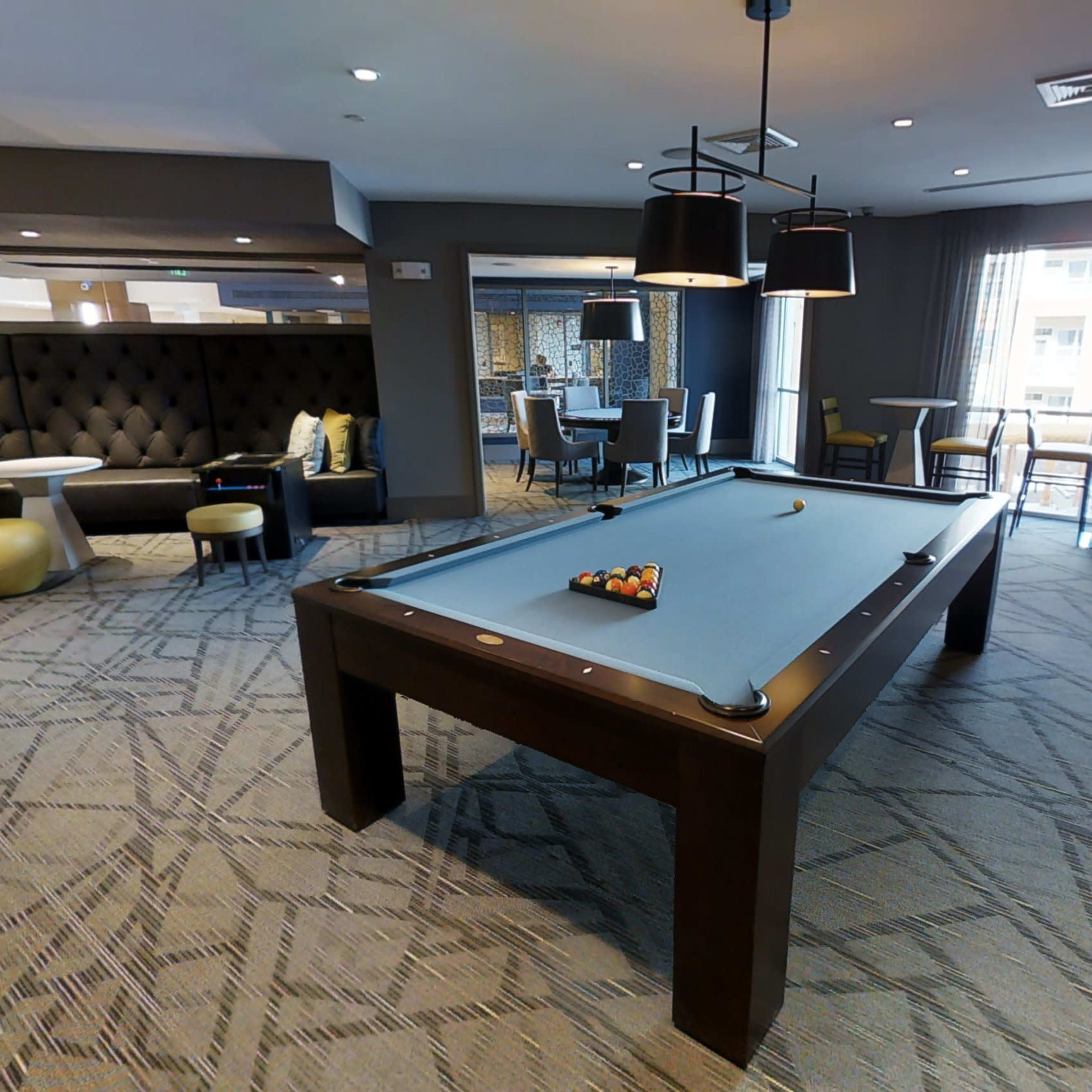 A modern lounge area featuring a pool table in the center, surrounded by seating and tables.