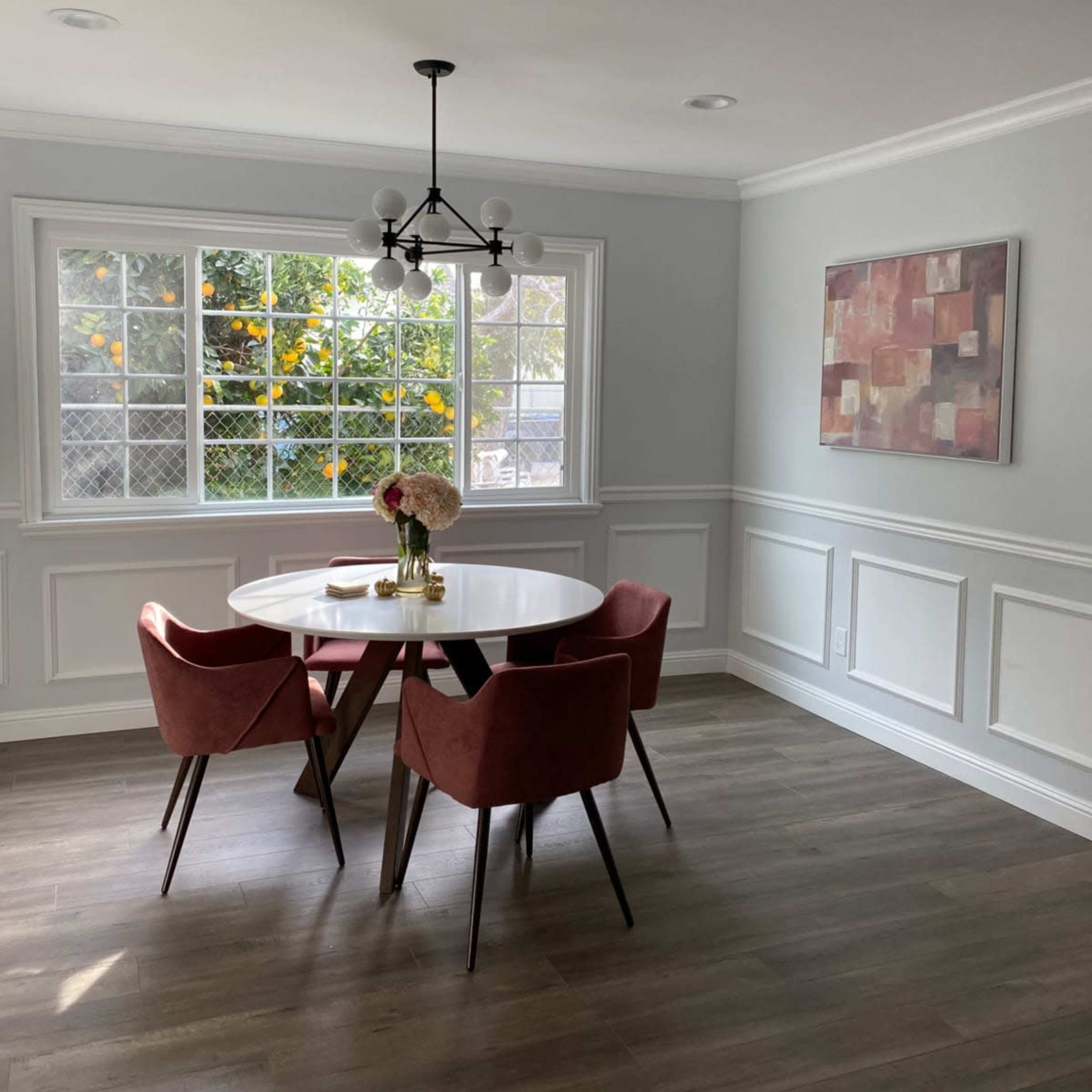 A modern dining area features a round white table surrounded by four pink chairs, next to a large window with greenery visible outside.