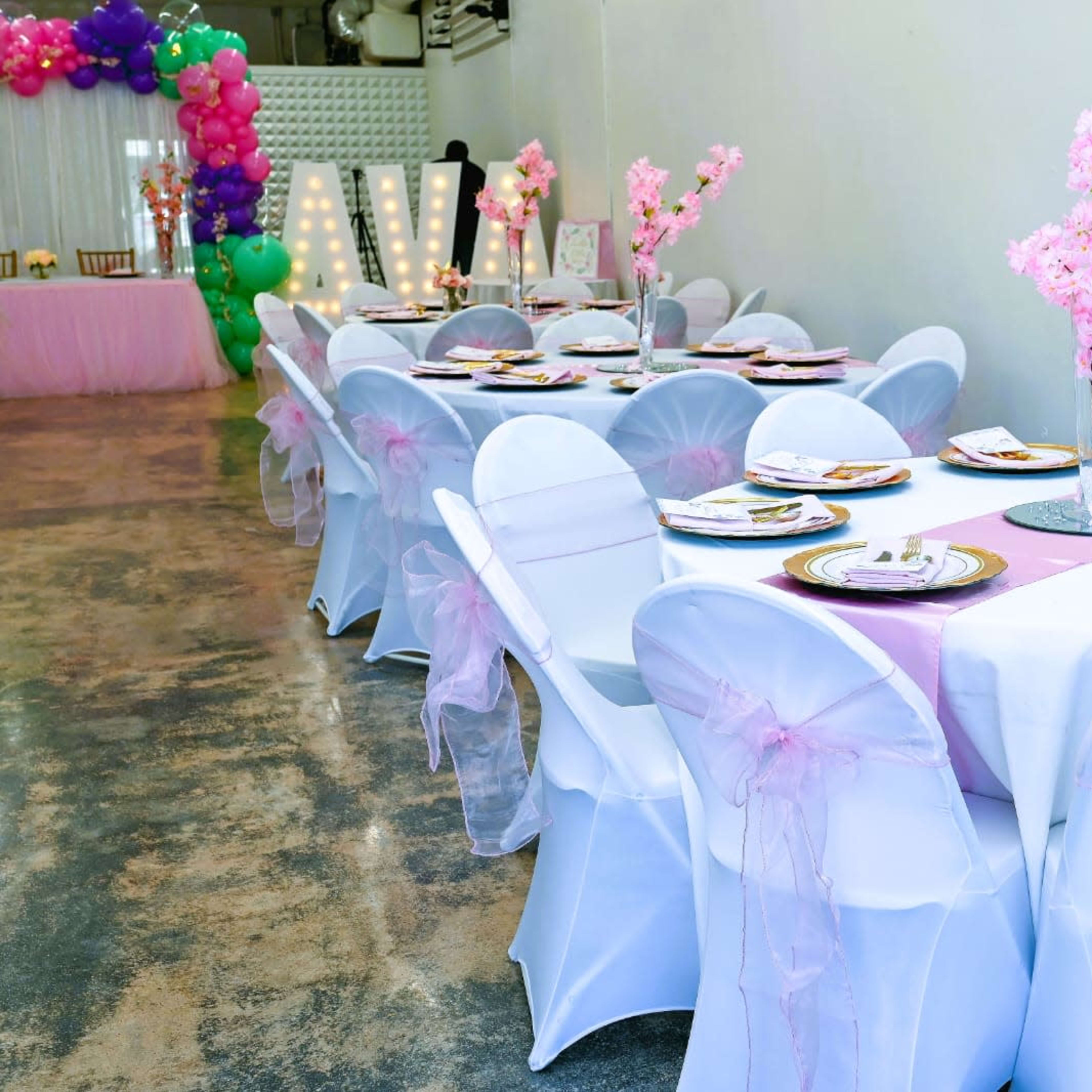 A banquet hall is set up with white tablecloths and chairs adorned with pink bows, featuring floral centerpieces on each table.