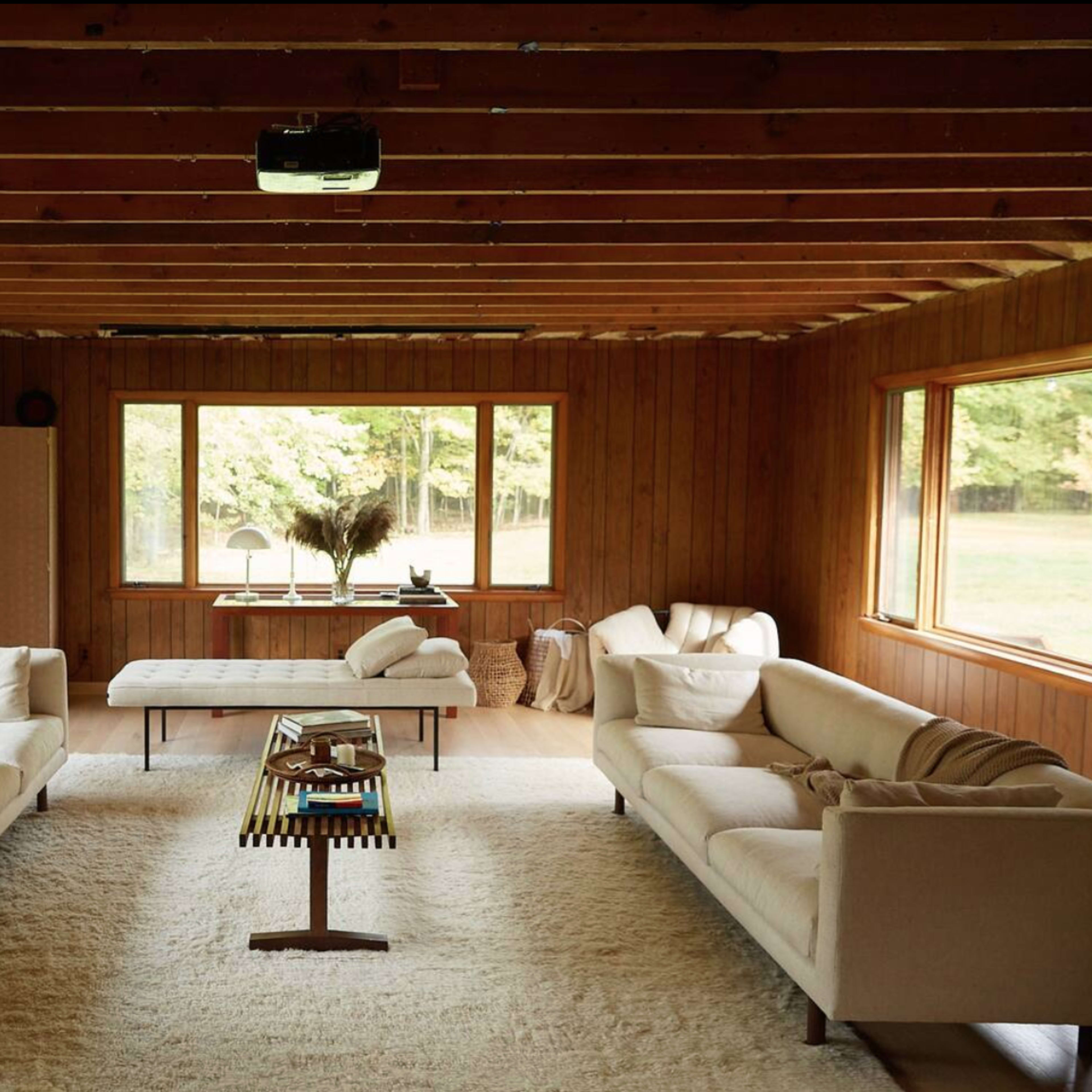 A cozy living room features two beige sofas facing a coffee table, with large windows overlooking a green landscape and wooden walls.