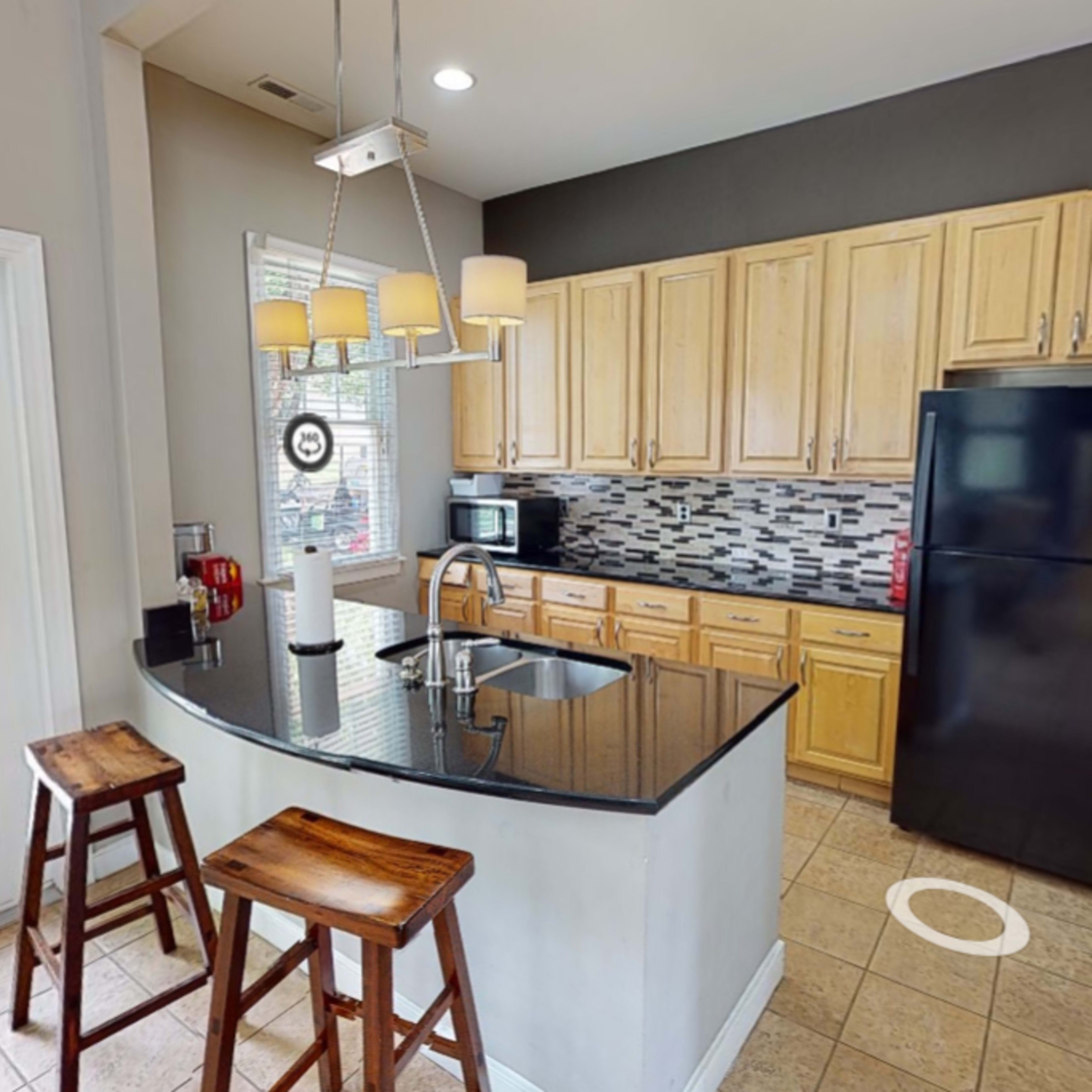 A modern kitchen featuring a curved granite countertop with bar stools, wooden cabinets, and a black refrigerator.