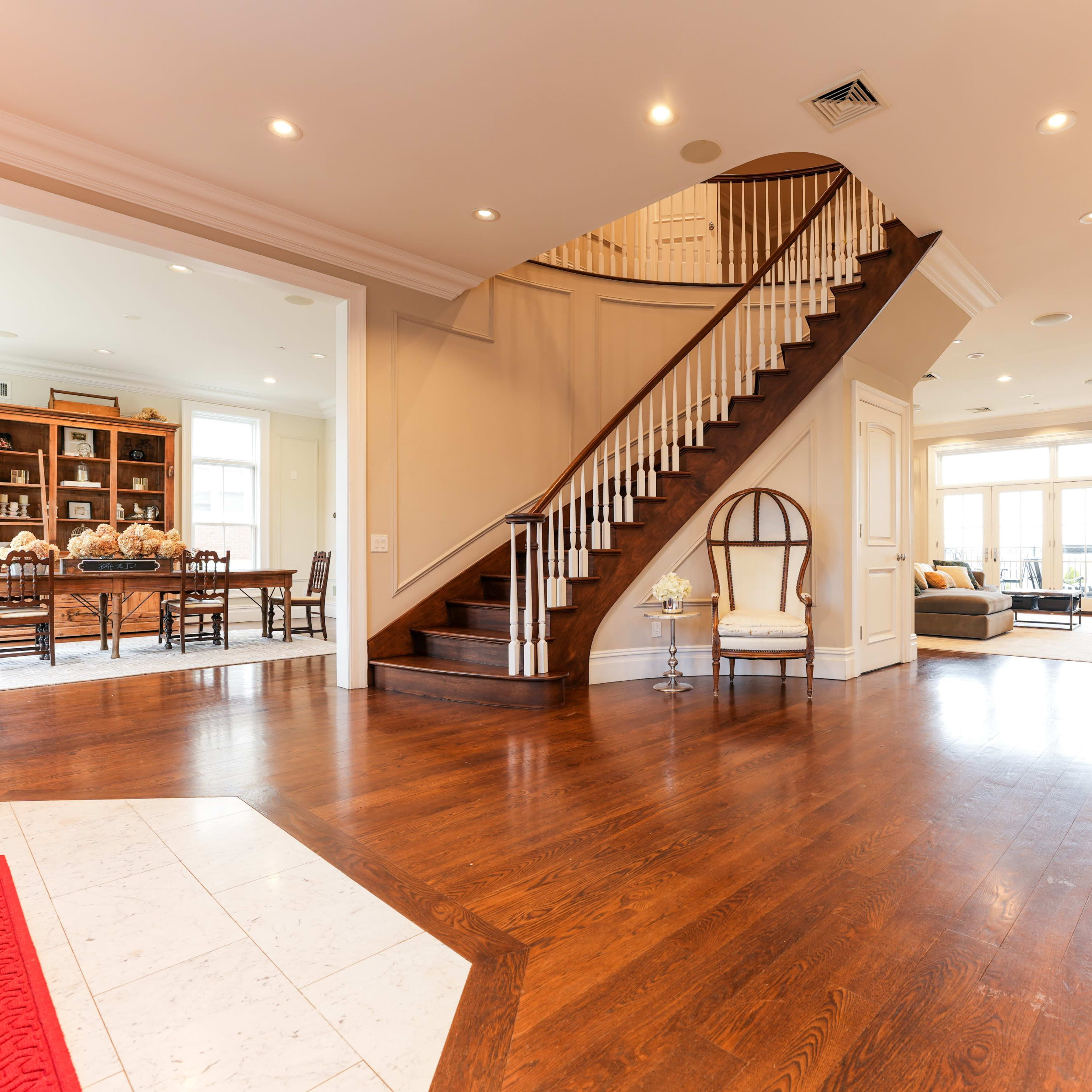 A spacious foyer with a wooden staircase leading to the upper level, adjoining a dining area and a living room with large windows.