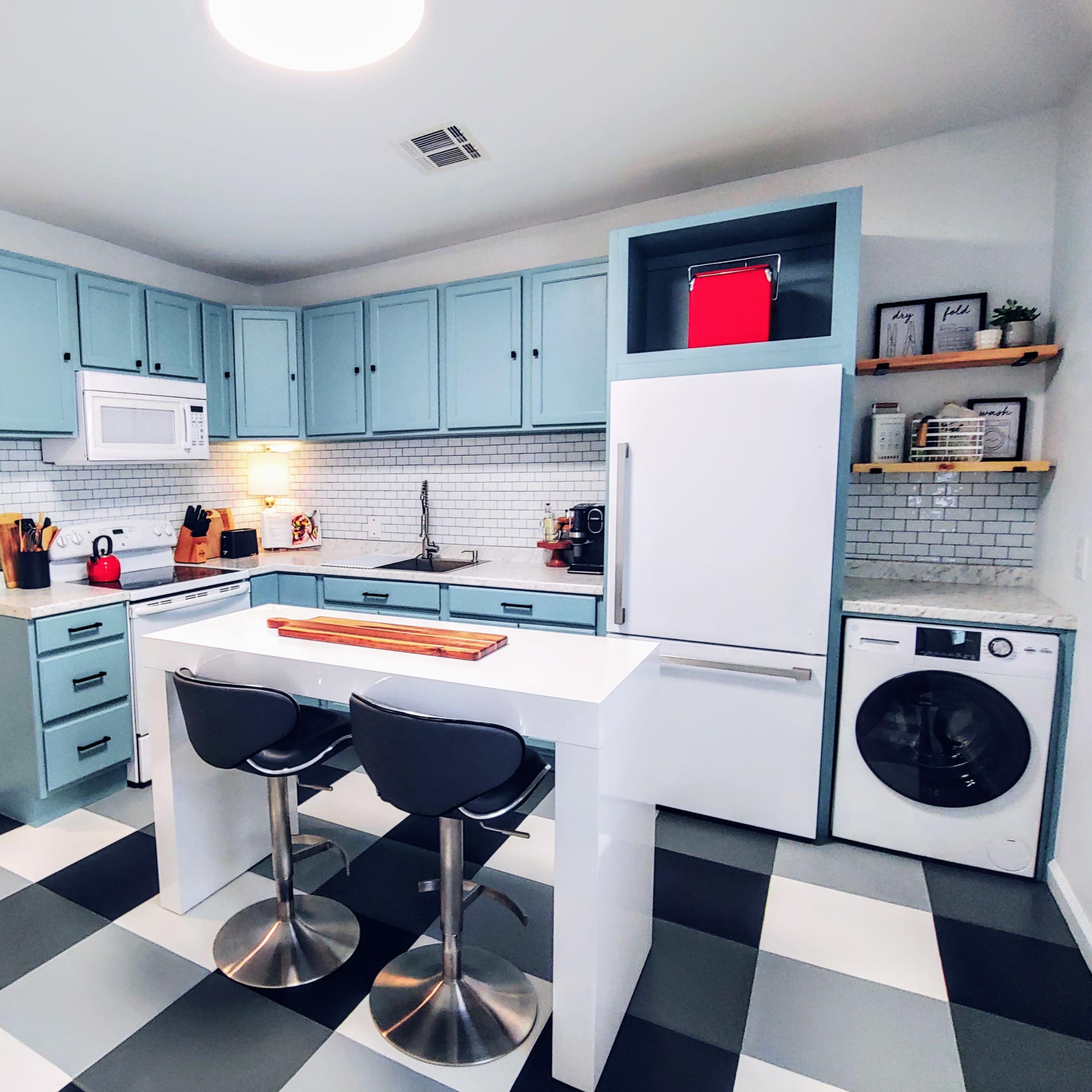 A modern kitchen with light blue cabinets, a white countertop, a washing machine, and a black and white checkered floor.