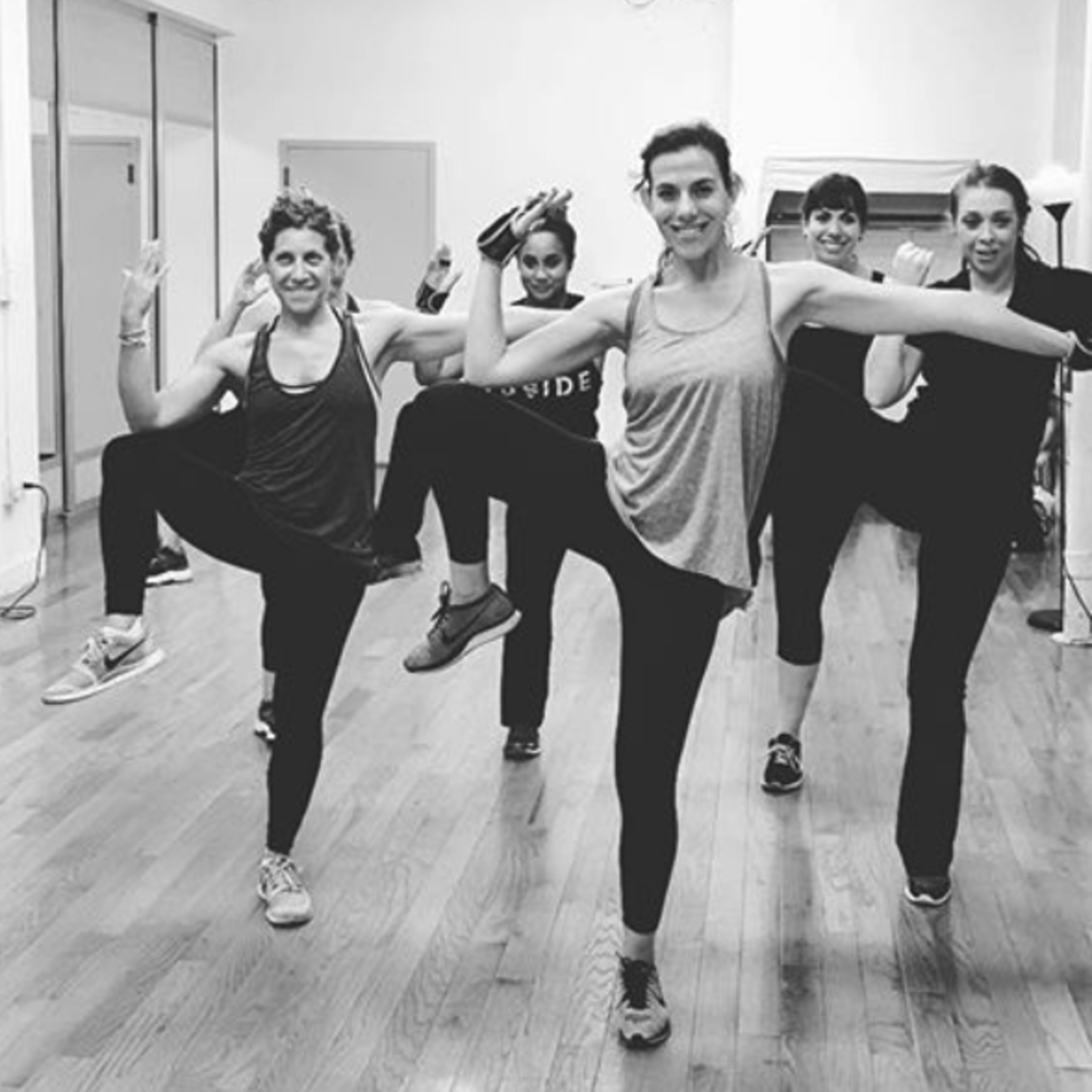 A group of six women is performing a dance routine in a studio with wooden floors and large windows.