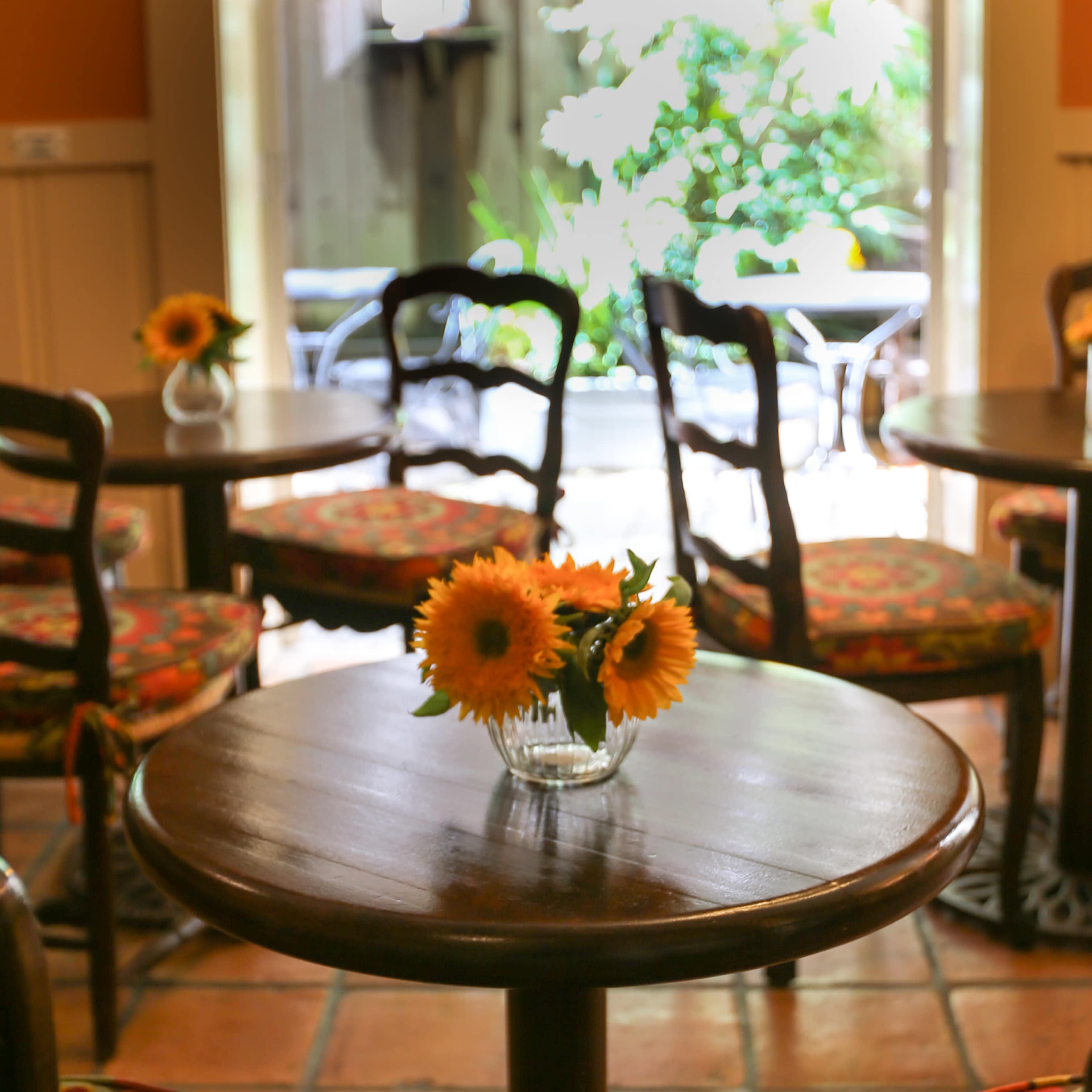A small wooden table with a vase of sunflowers is surrounded by chairs in a cafe with an outdoor view.