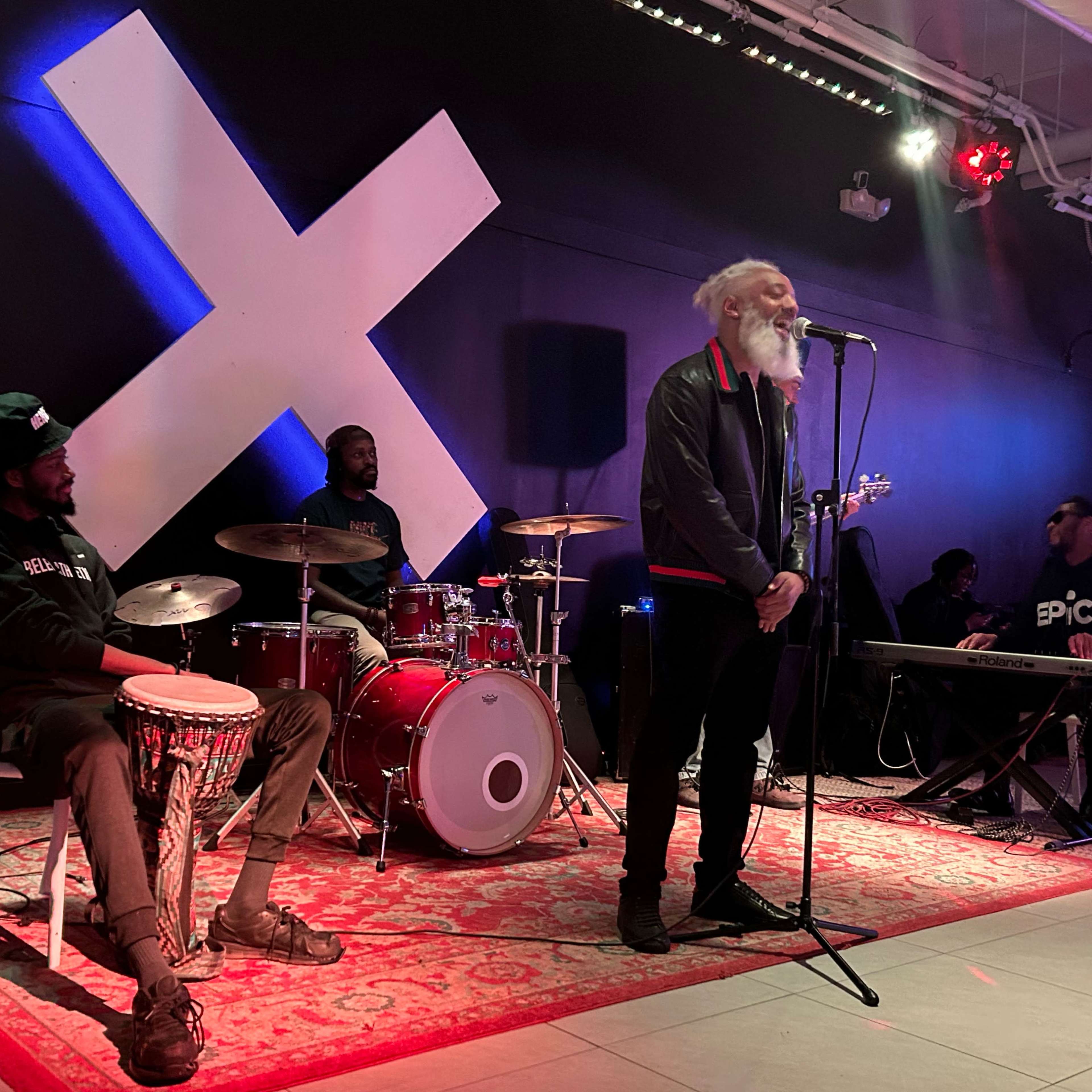 A live music performance features a singer speaking into a microphone while seated musicians play drums and percussion.