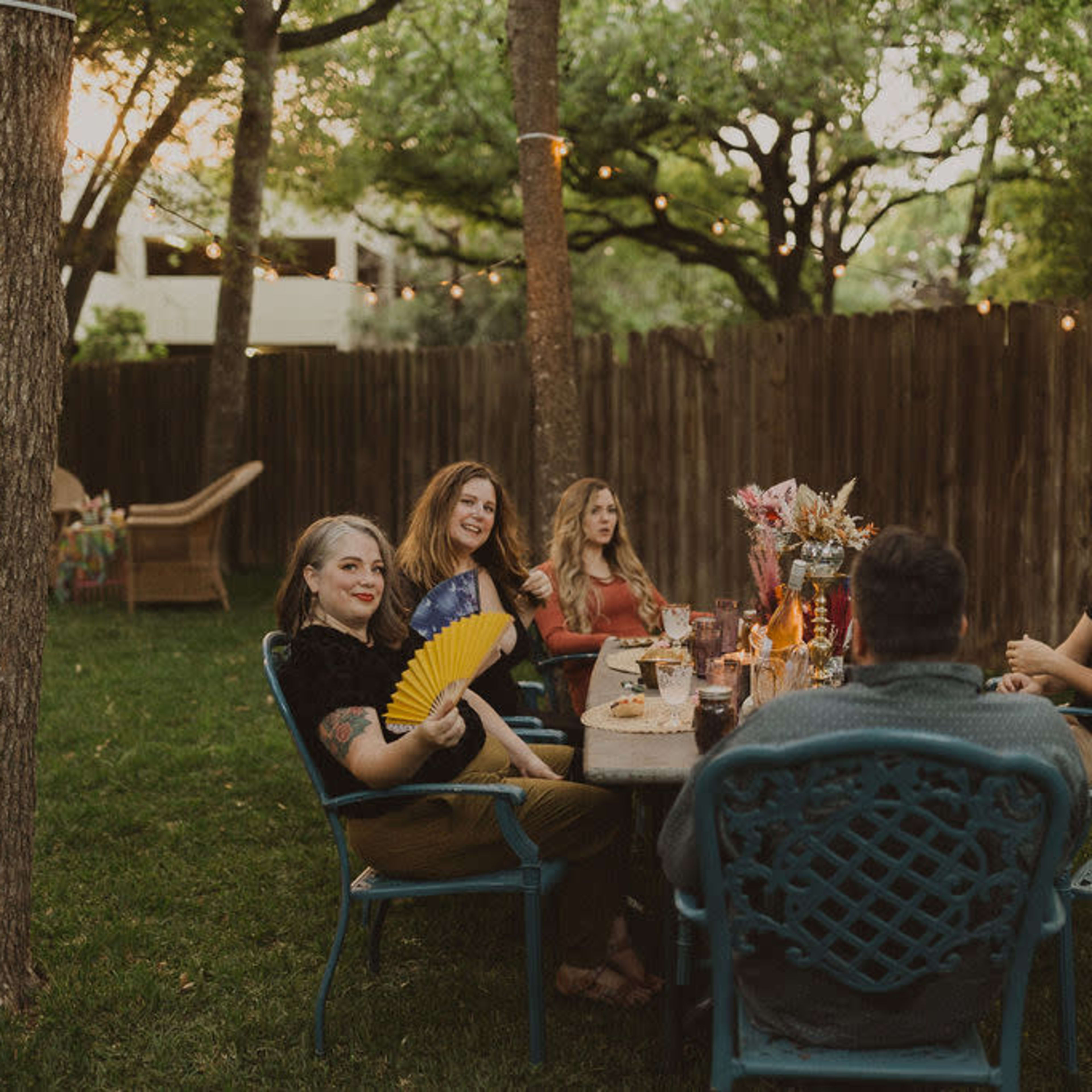 A group of five people sits around a table in a backyard, enjoying drinks and conversation amidst trees and string lights.