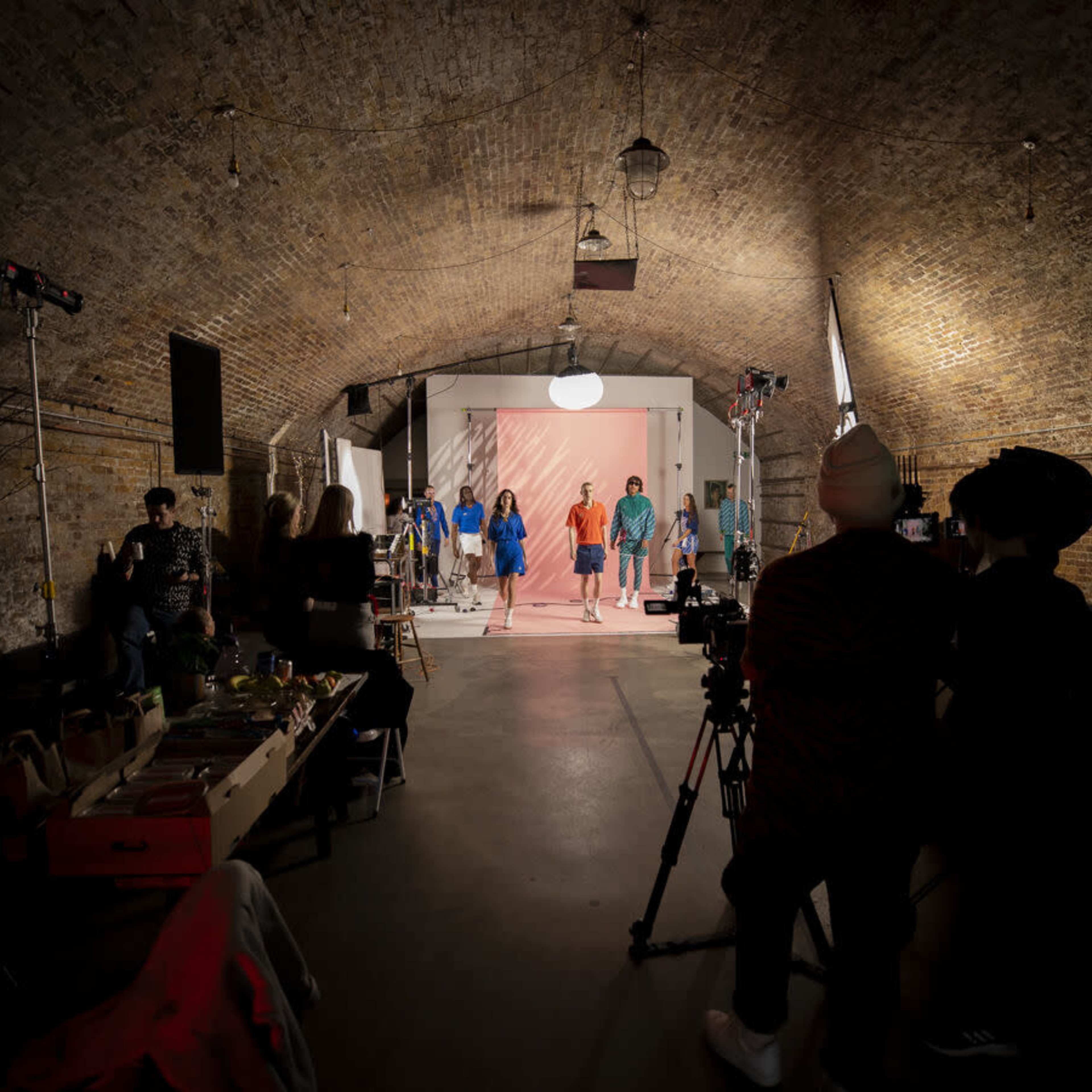 A group of models walks down a runway in a brick-walled space filled with cameras and lighting equipment.