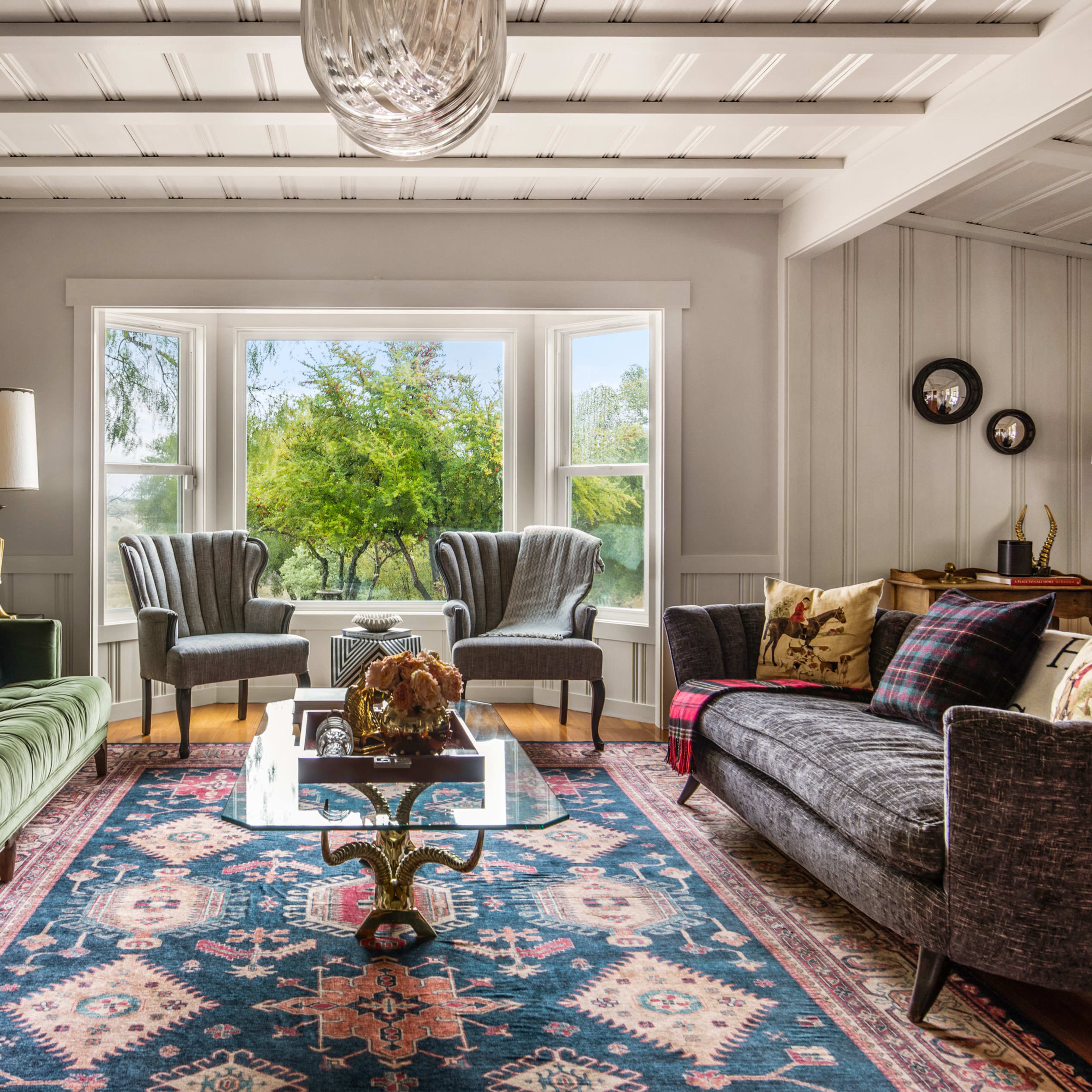 A living room features green and gray sofas, a glass coffee table, and large windows overlooking a lush landscape.