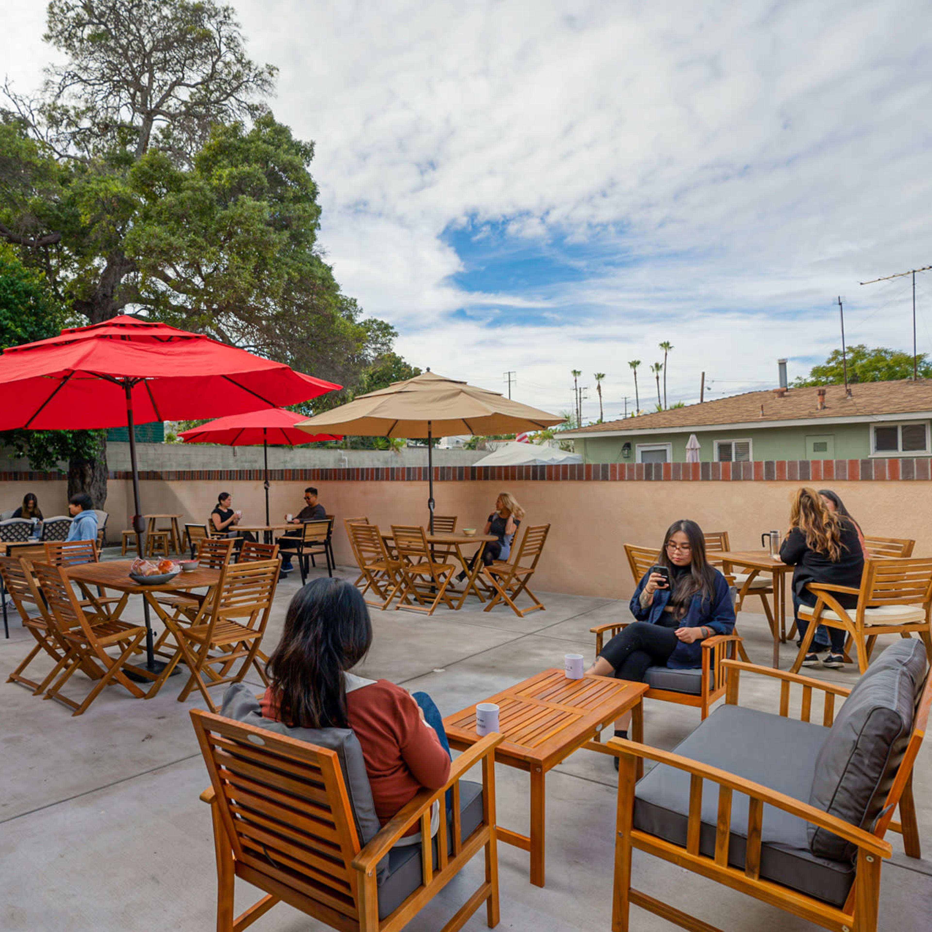 A patio area with several groups of people seated at wooden tables beneath red and beige umbrellas.