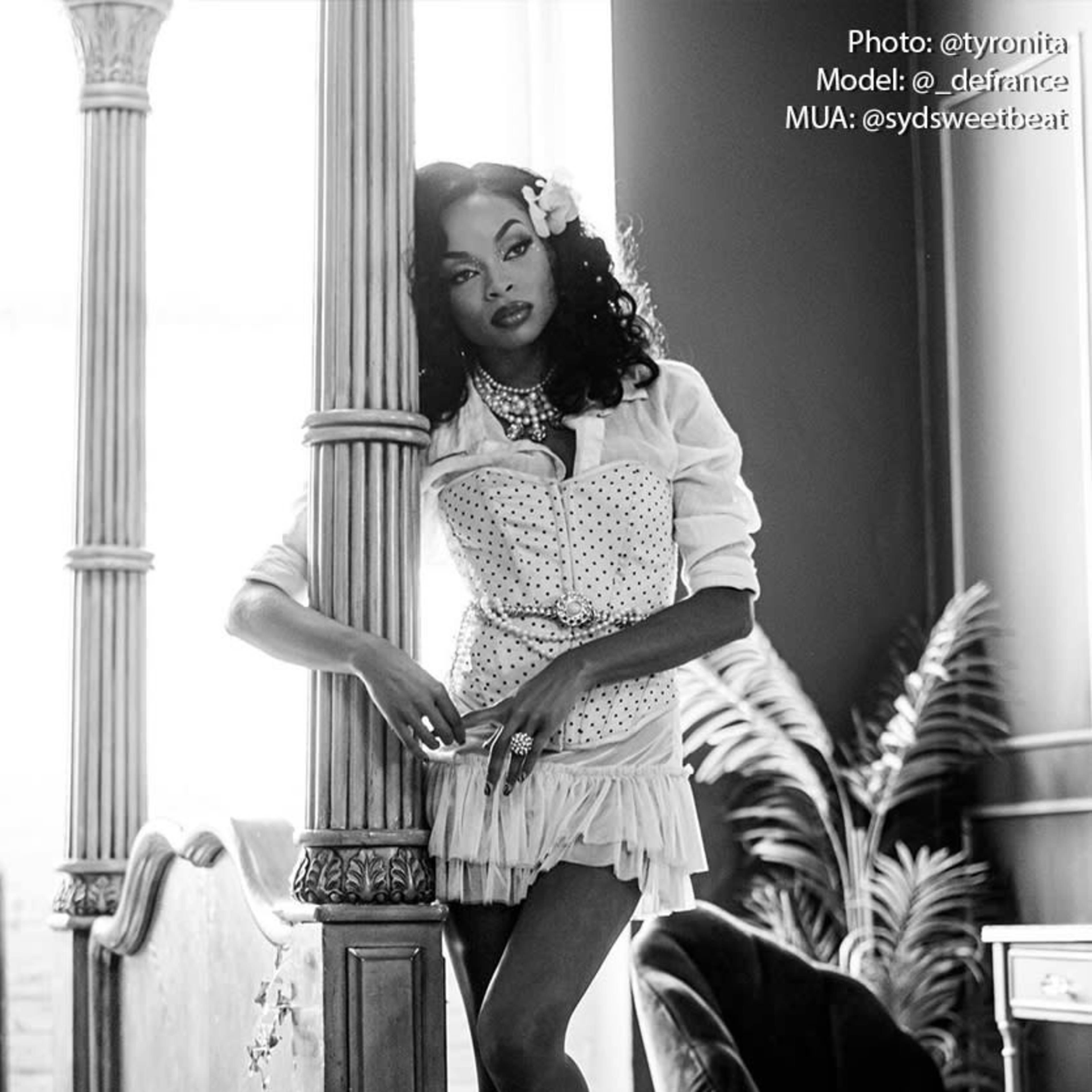 A model poses elegantly by a vintage bed, wearing a white corset and a flower in her hair, with a backdrop of tall windows and greenery.