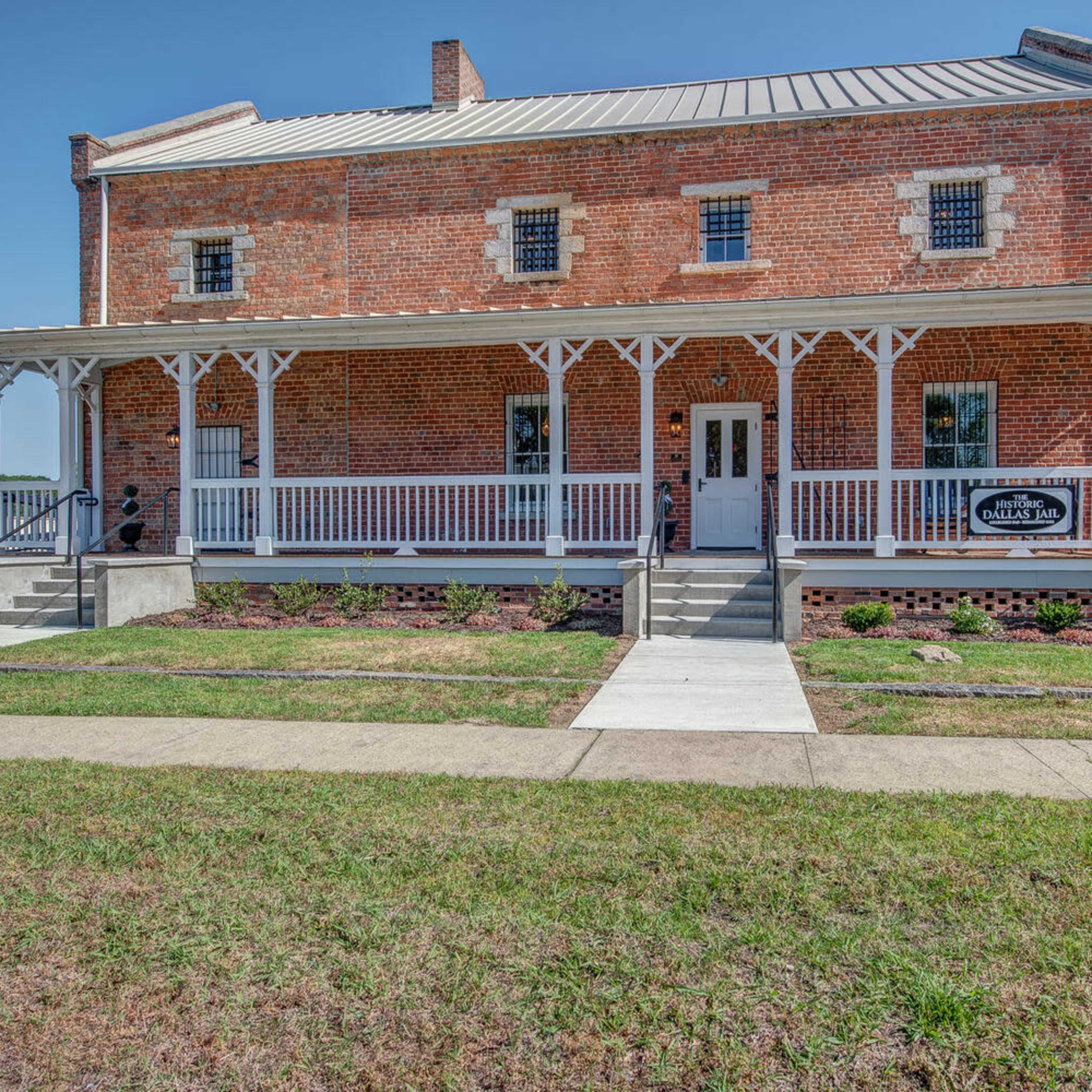 A two-story brick building with a front porch and white railings, located on a grassy area with a sidewalk leading to the entrance.