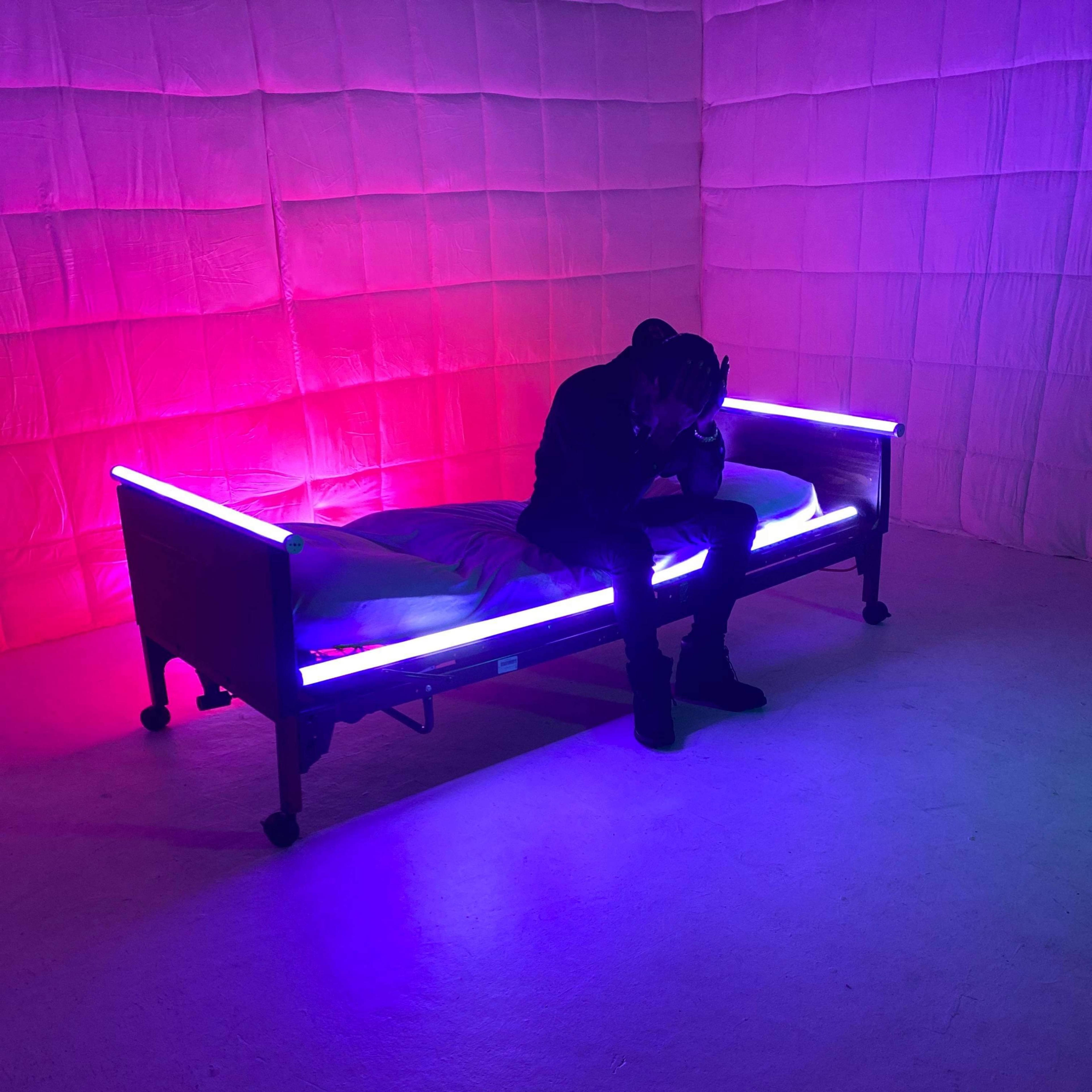 A person sits on a bed with neon lighting in a dimly lit room.
