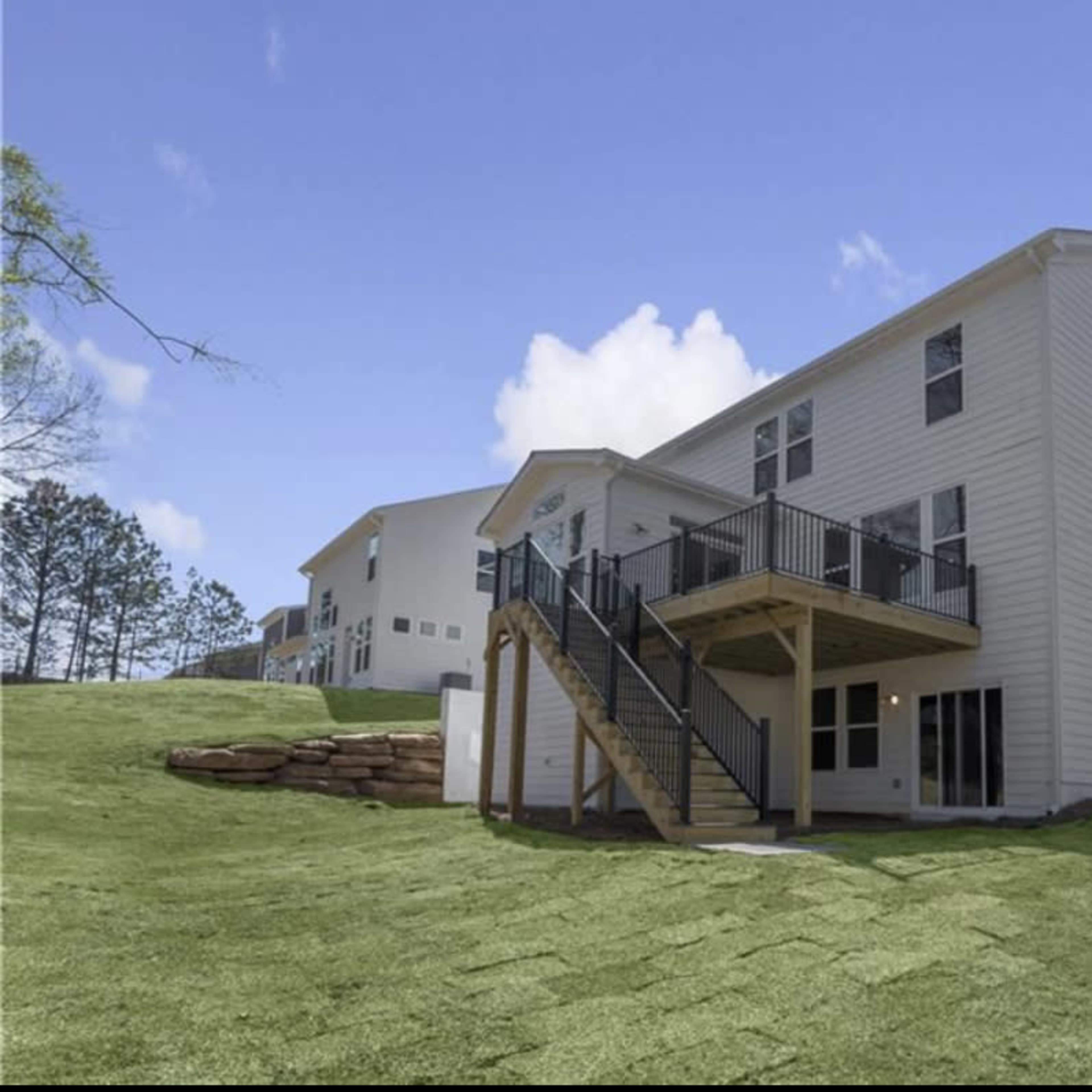 A two-story house with a wooden deck, located on a grassy hillside surrounded by trees.