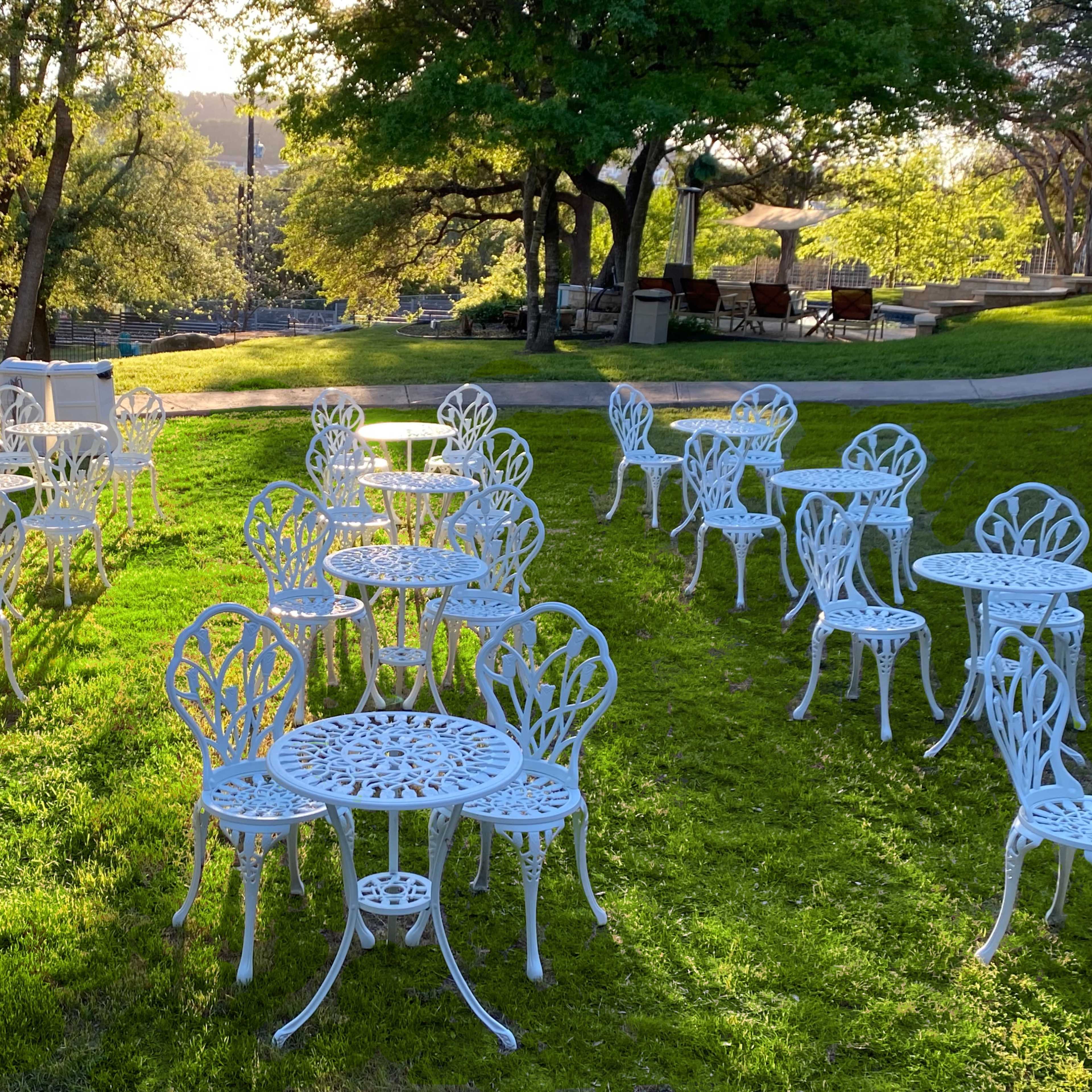 A grassy area is adorned with white metal tables and chairs arranged in clusters under the soft light of the setting sun.