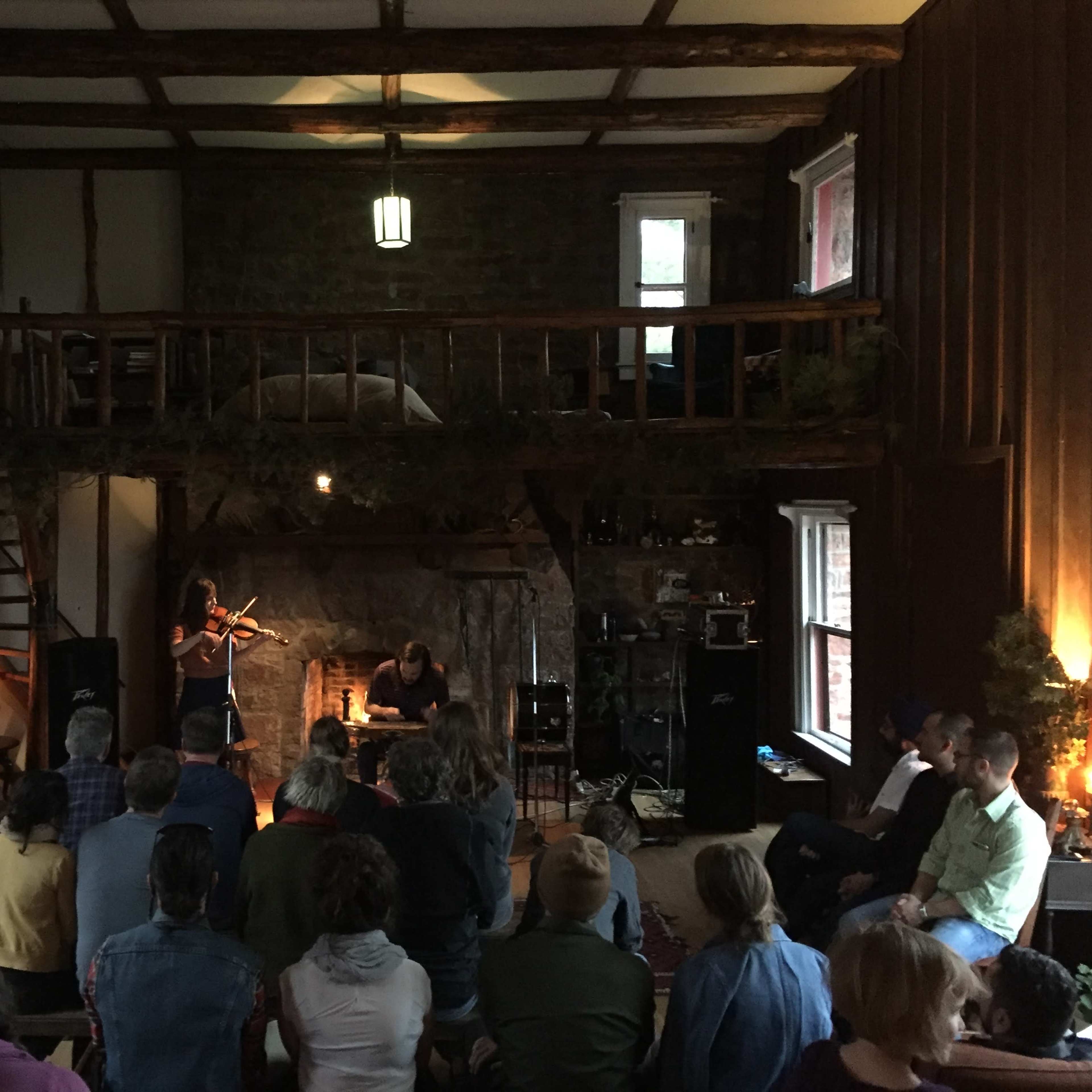 A group of people sit in a cozy room with a stone fireplace, watching a performer play the violin while another musician sits nearby.