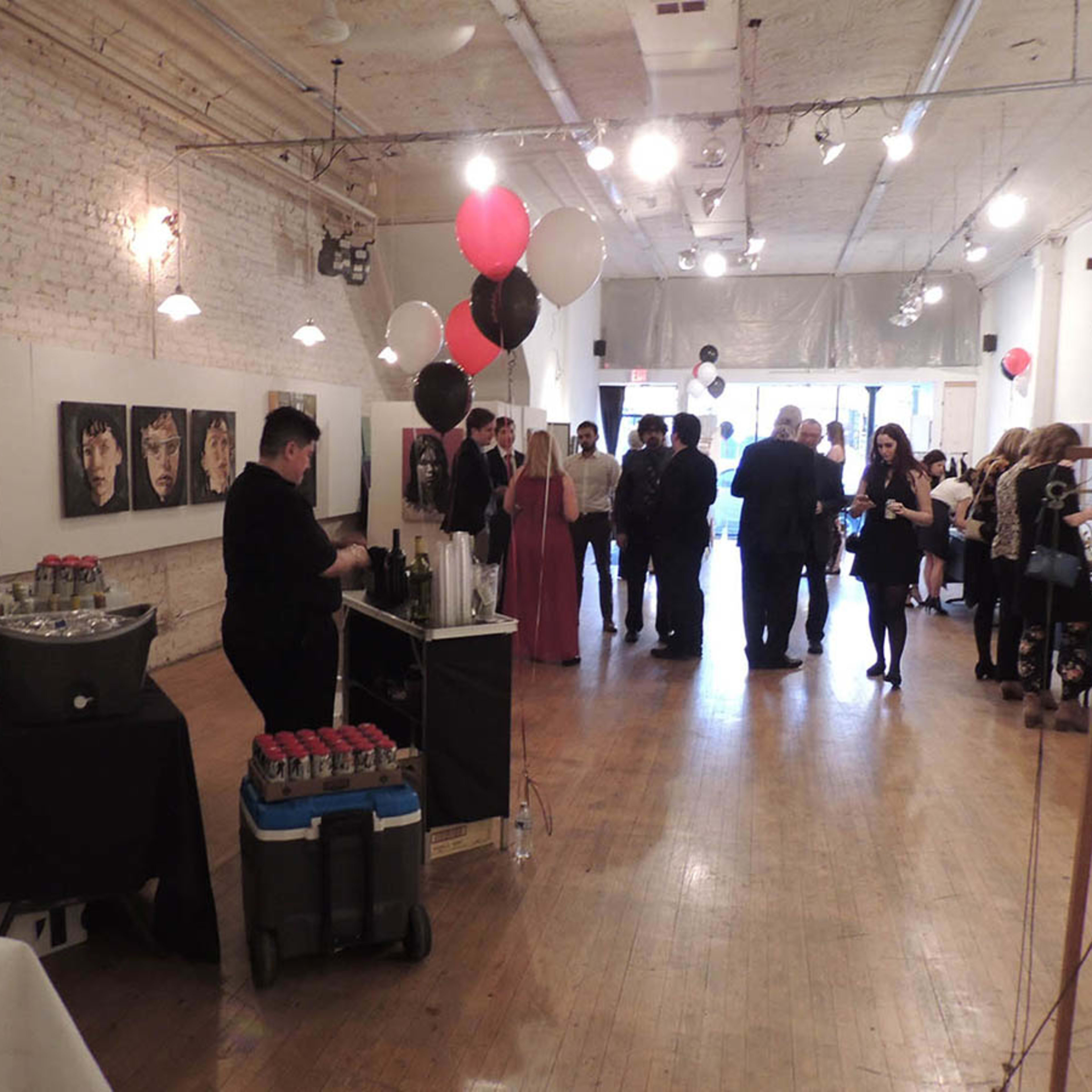 A spacious art gallery with guests mingling, a black table with drinks on one side, and artwork hanging on the walls.