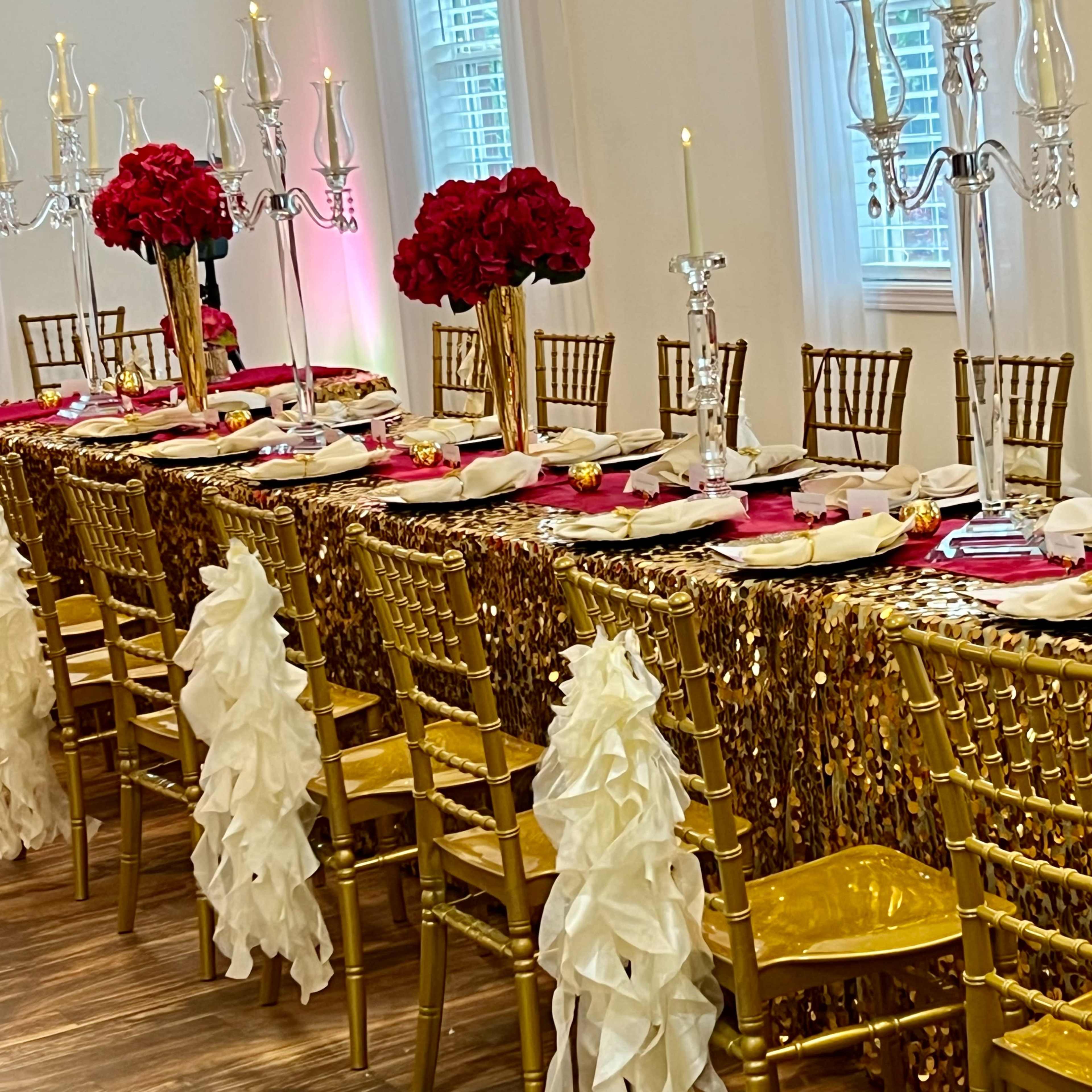 A long dining table is set with gold and white decor, featuring red floral centerpieces and elegant tableware.