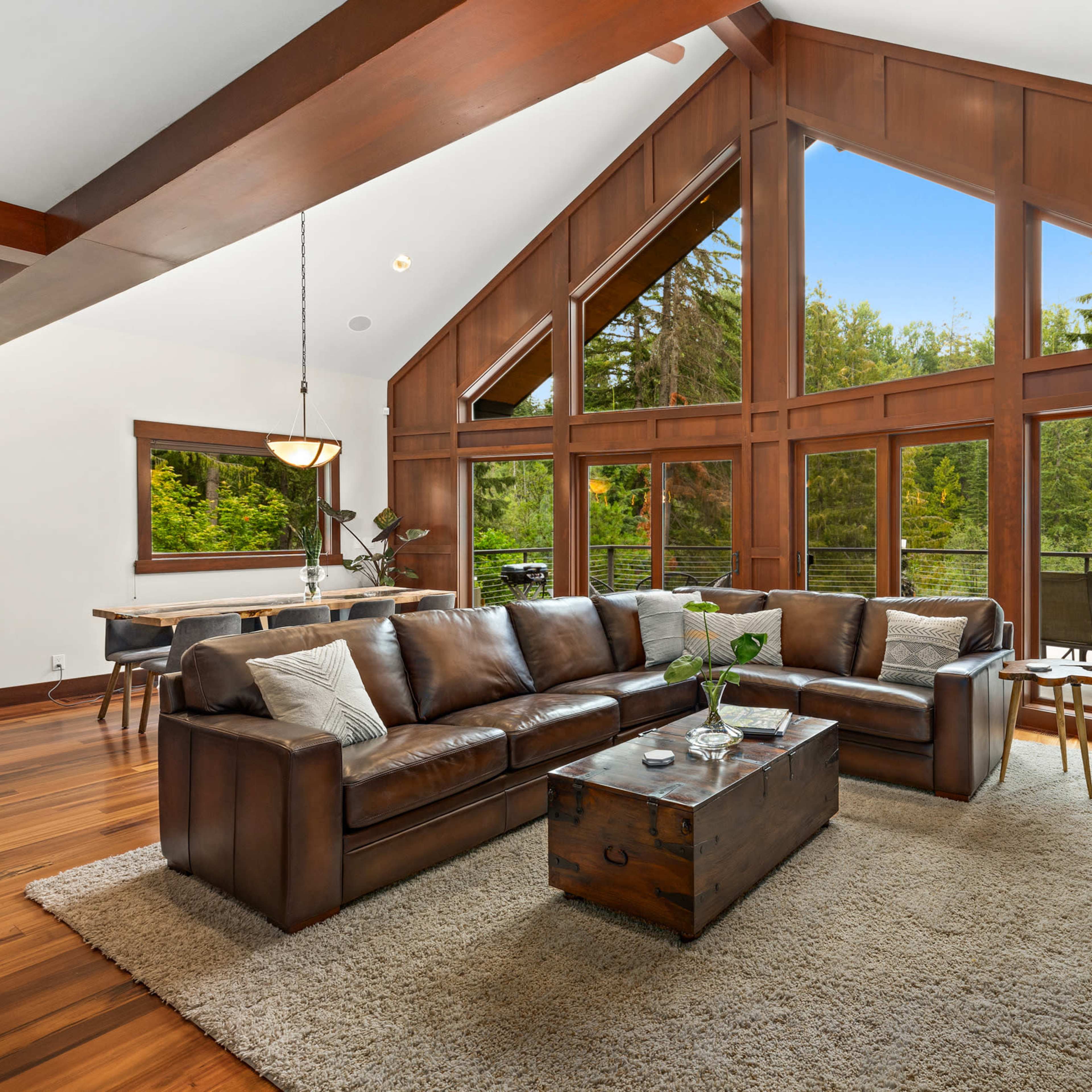 A spacious living room features a large leather sectional sofa, a wooden coffee table, and expansive windows that offer views of a forested outdoor setting.