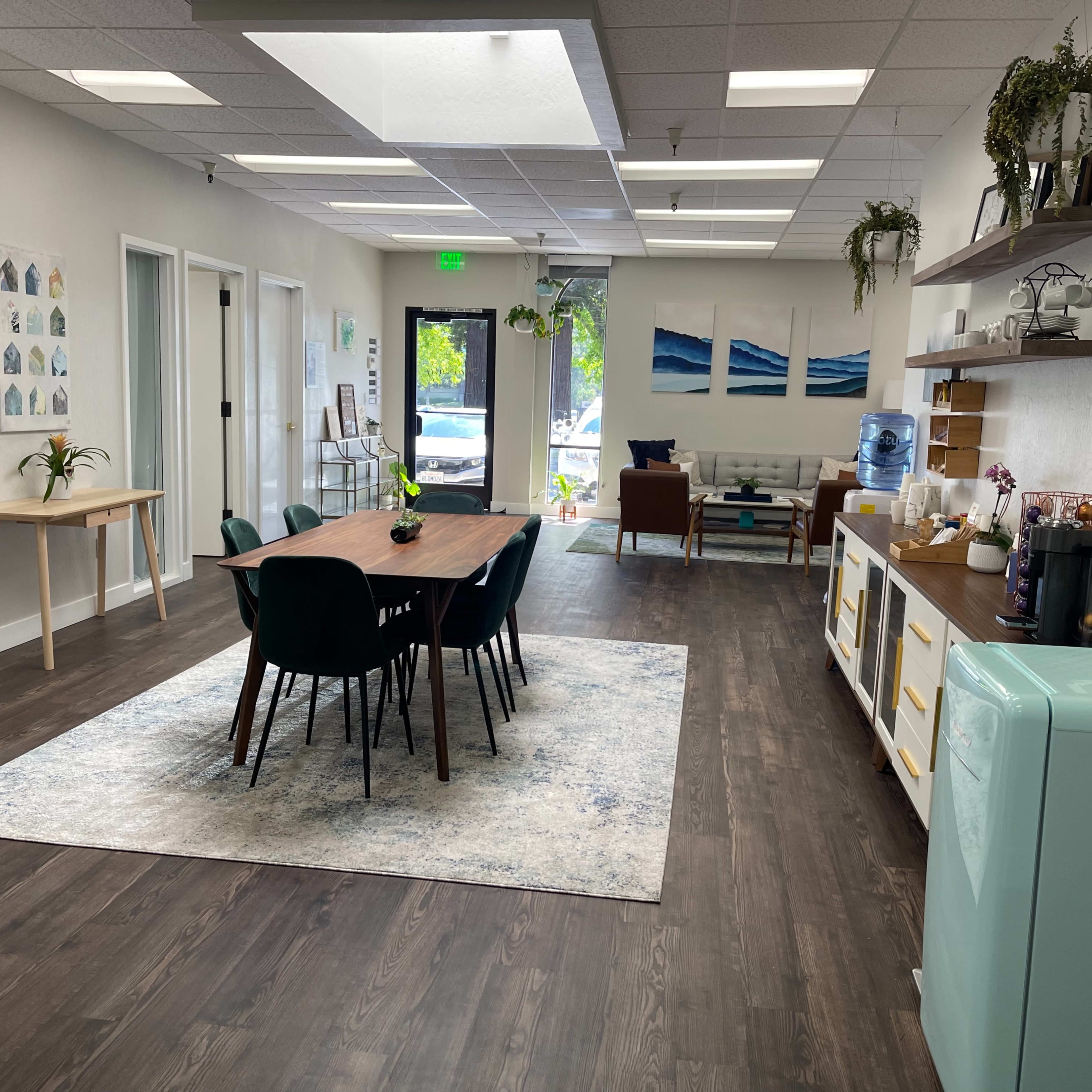 A modern office space with a dining table surrounded by chairs, a kitchenette, and a reception area featuring decorative plants and artwork.