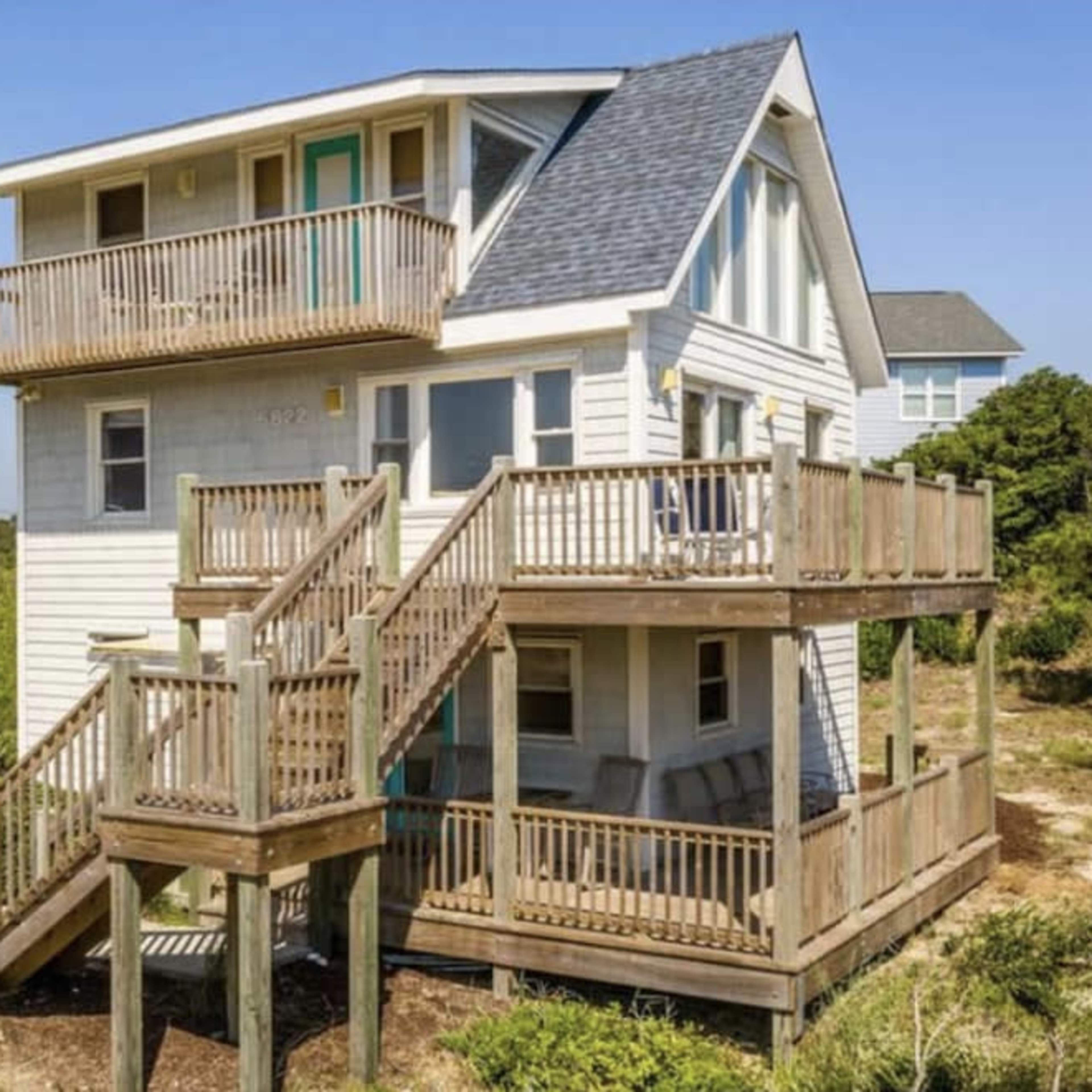 A two-story beach house with multiple balconies and wooden decks is situated among sandy vegetation.