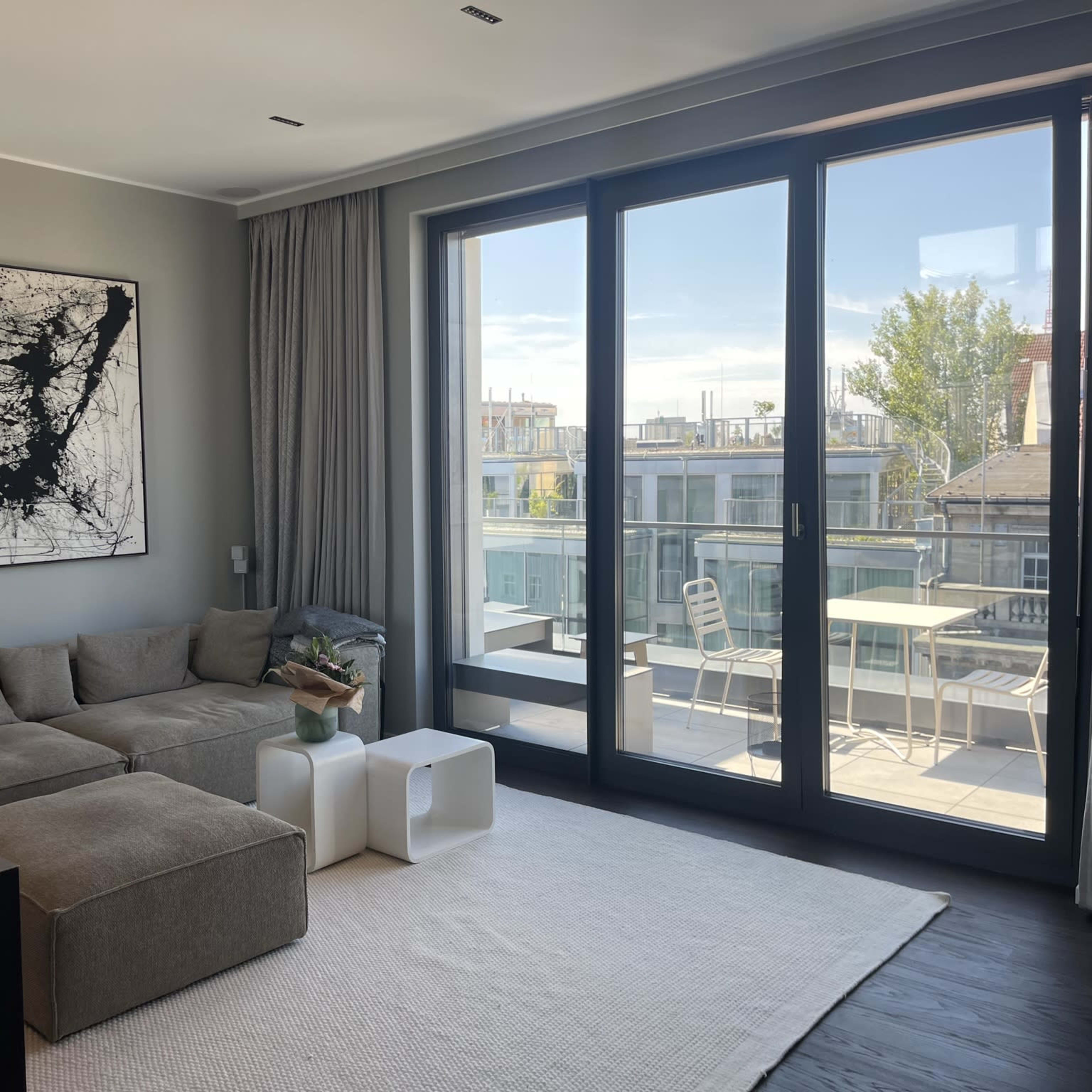 A modern living room features a large sectional sofa, a minimalist coffee table, and a large window that opens to a balcony with outdoor seating.