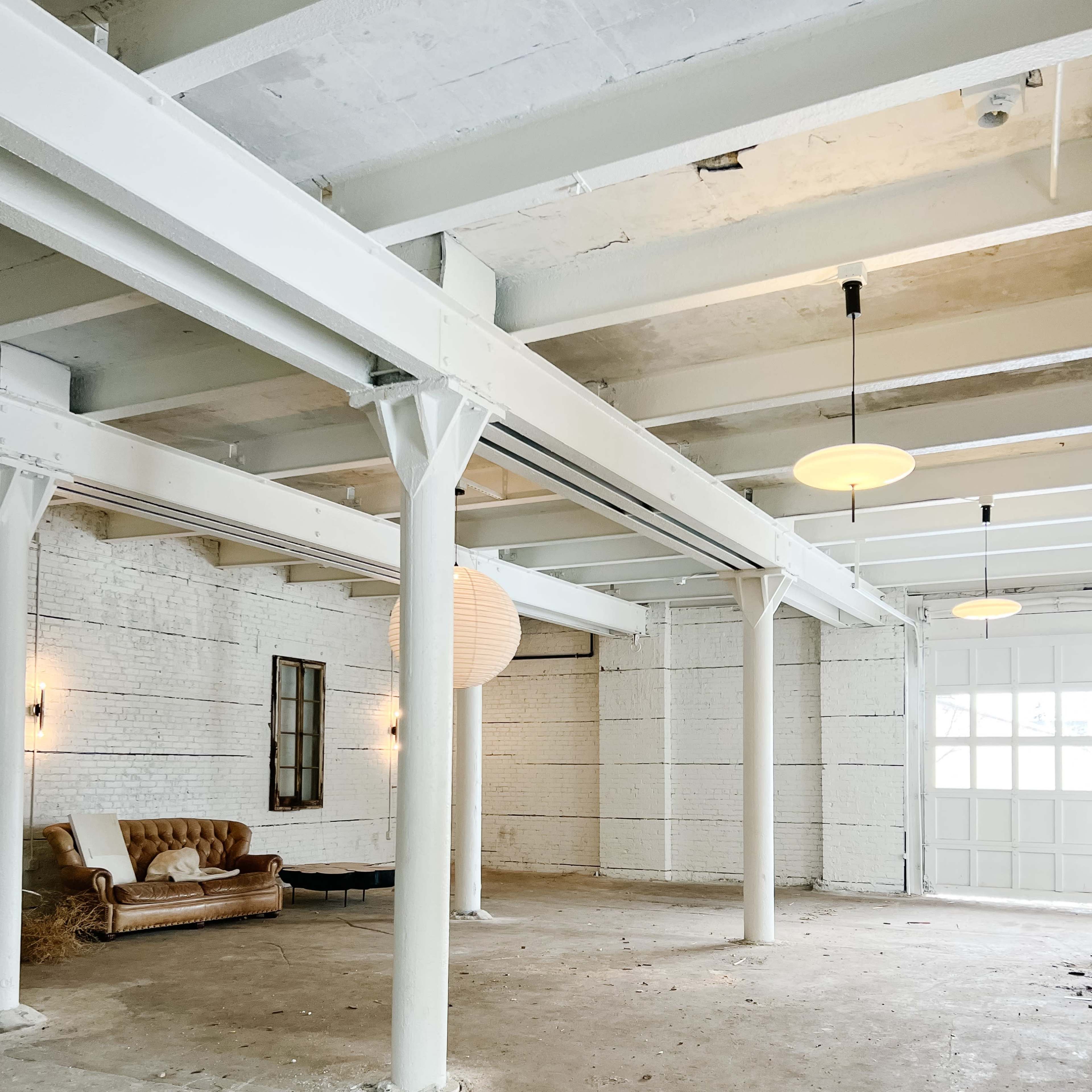 An empty, industrial-style interior with white walls, exposed beams, and a brown leather couch against one of the walls.