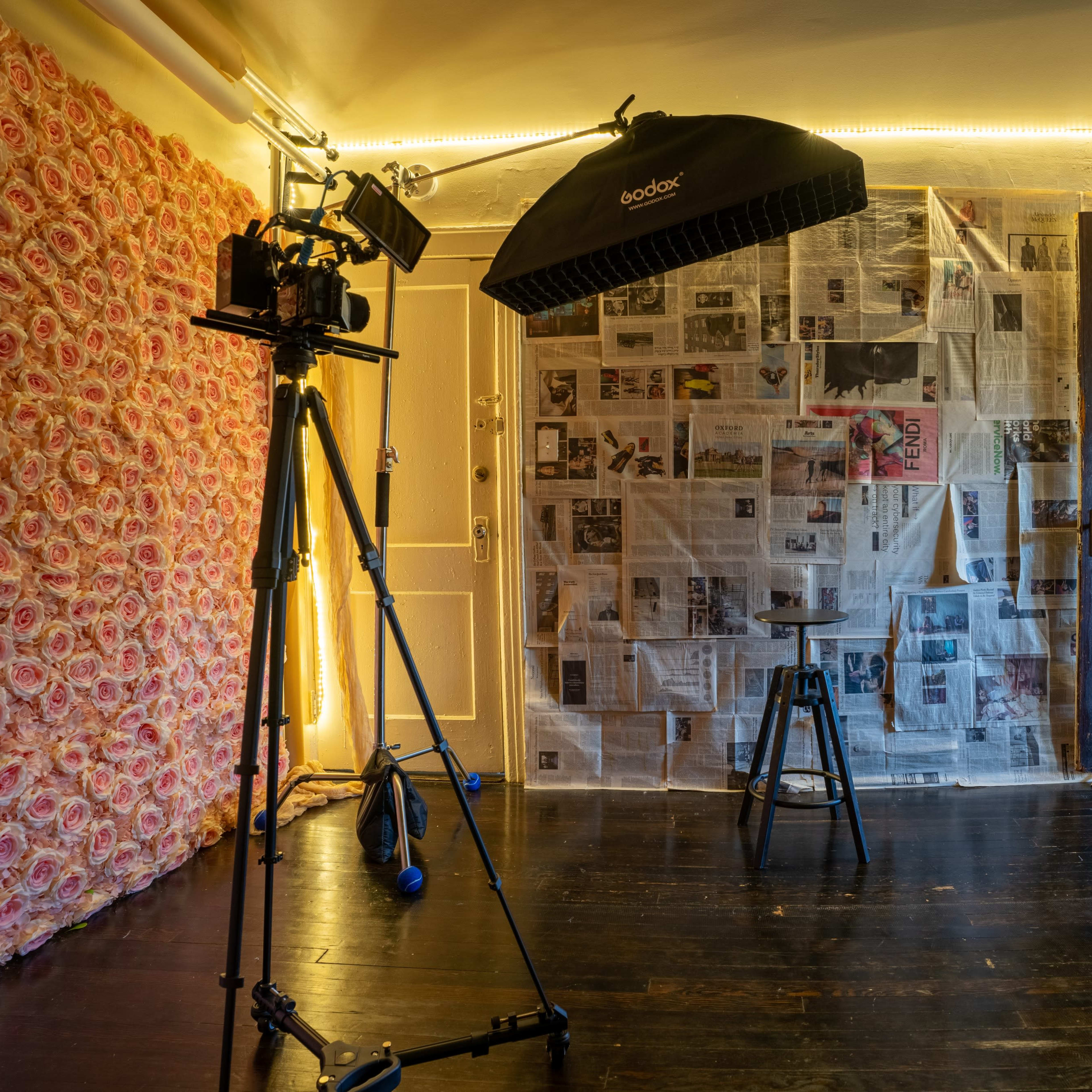 A photography studio with a camera on a tripod, a stool, and a backdrop made of newspapers, surrounded by a floral wall and illuminated by string lights.