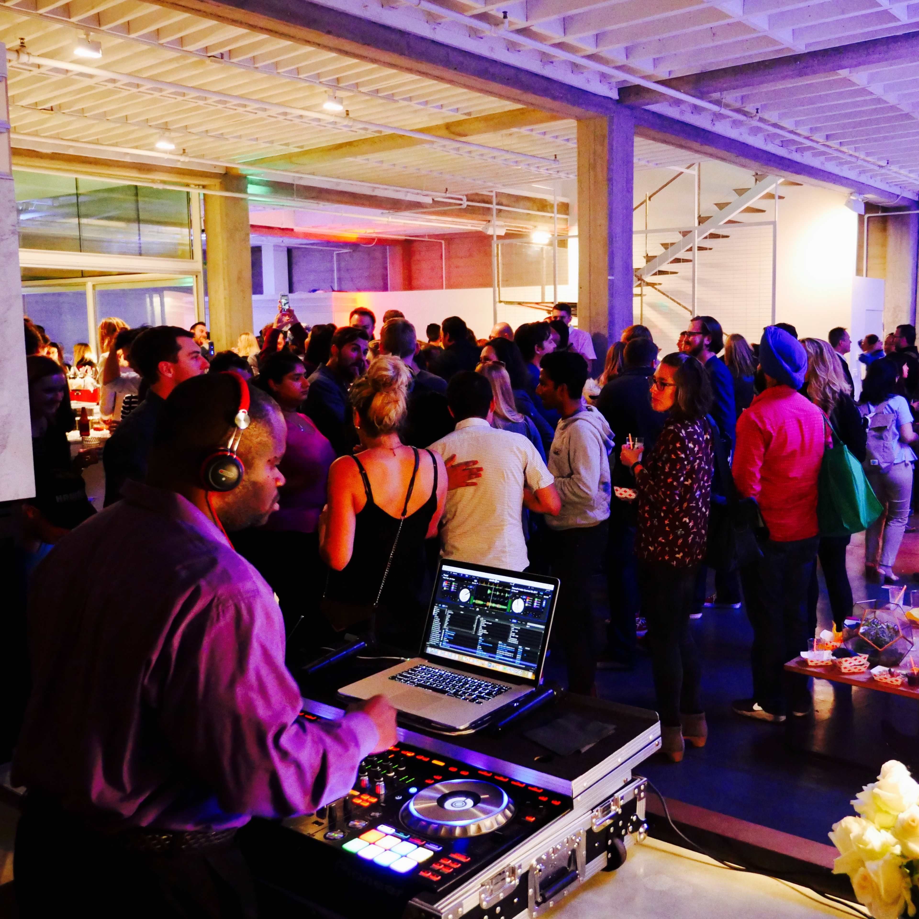 A DJ controls the music at a lively gathering filled with guests mingling in a well-lit space.