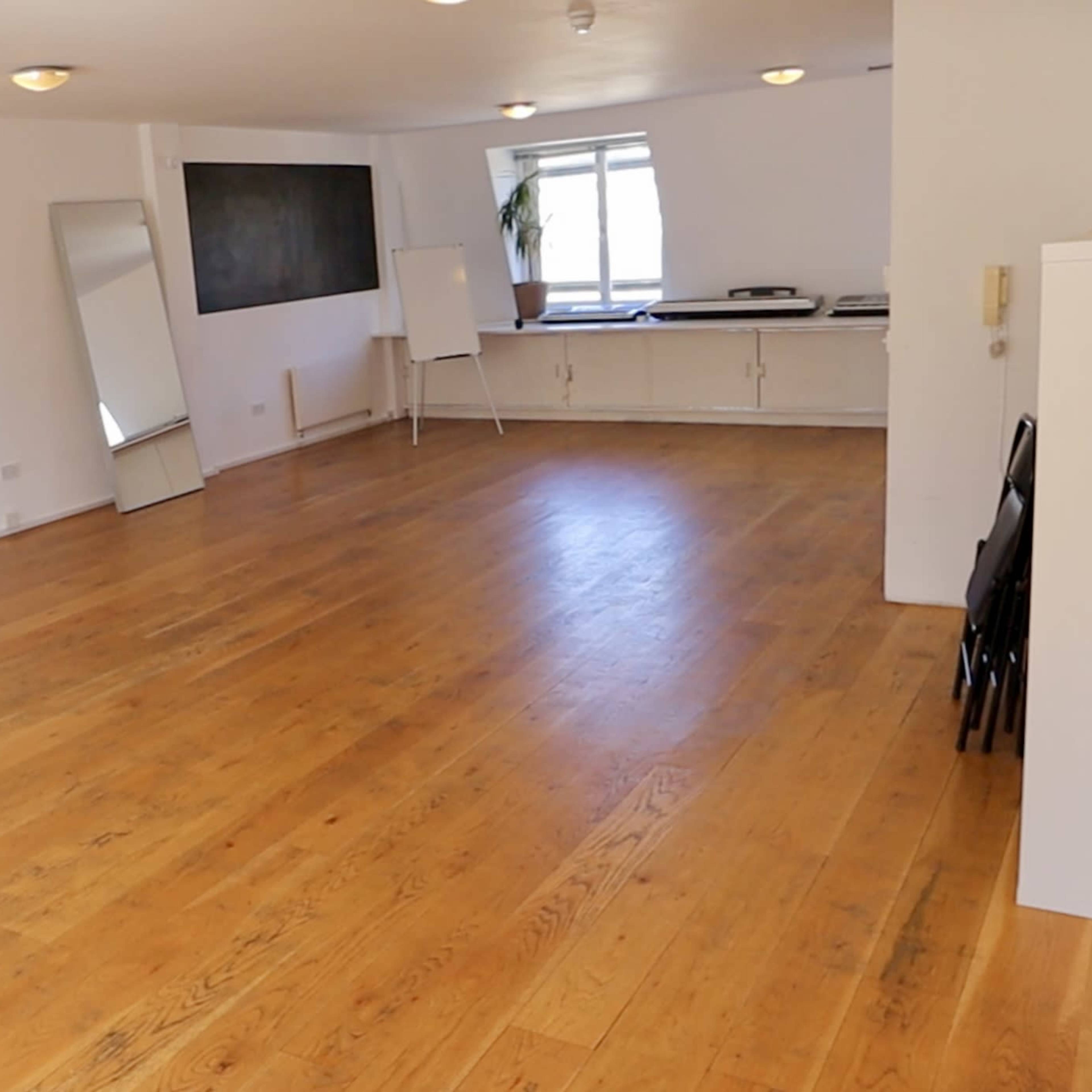 A spacious, empty room with wooden flooring, large windows, a mirror, and a whiteboard at one end.