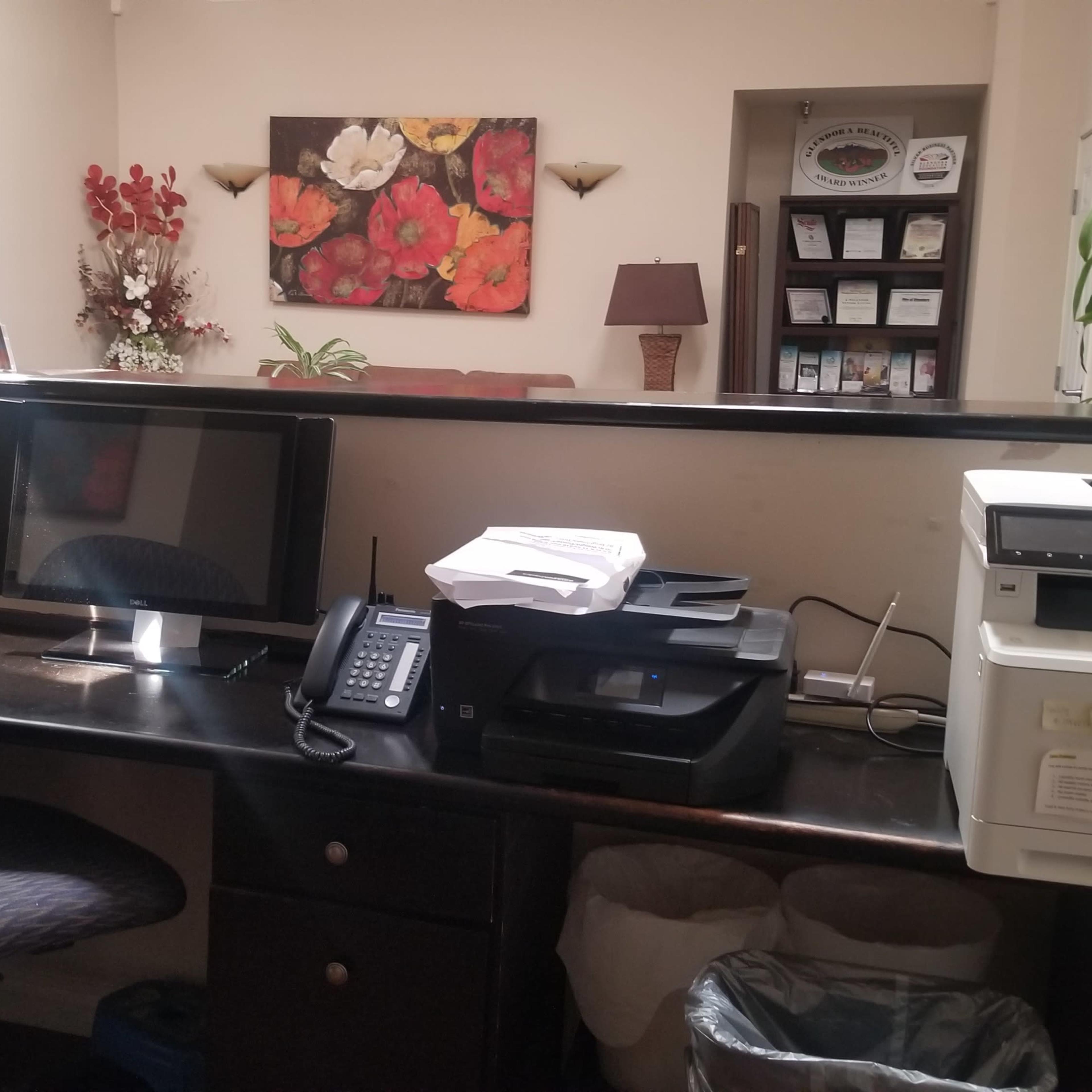 An office reception area with a desk featuring a computer monitor, a phone, a printer, and decorative flowers on the wall.