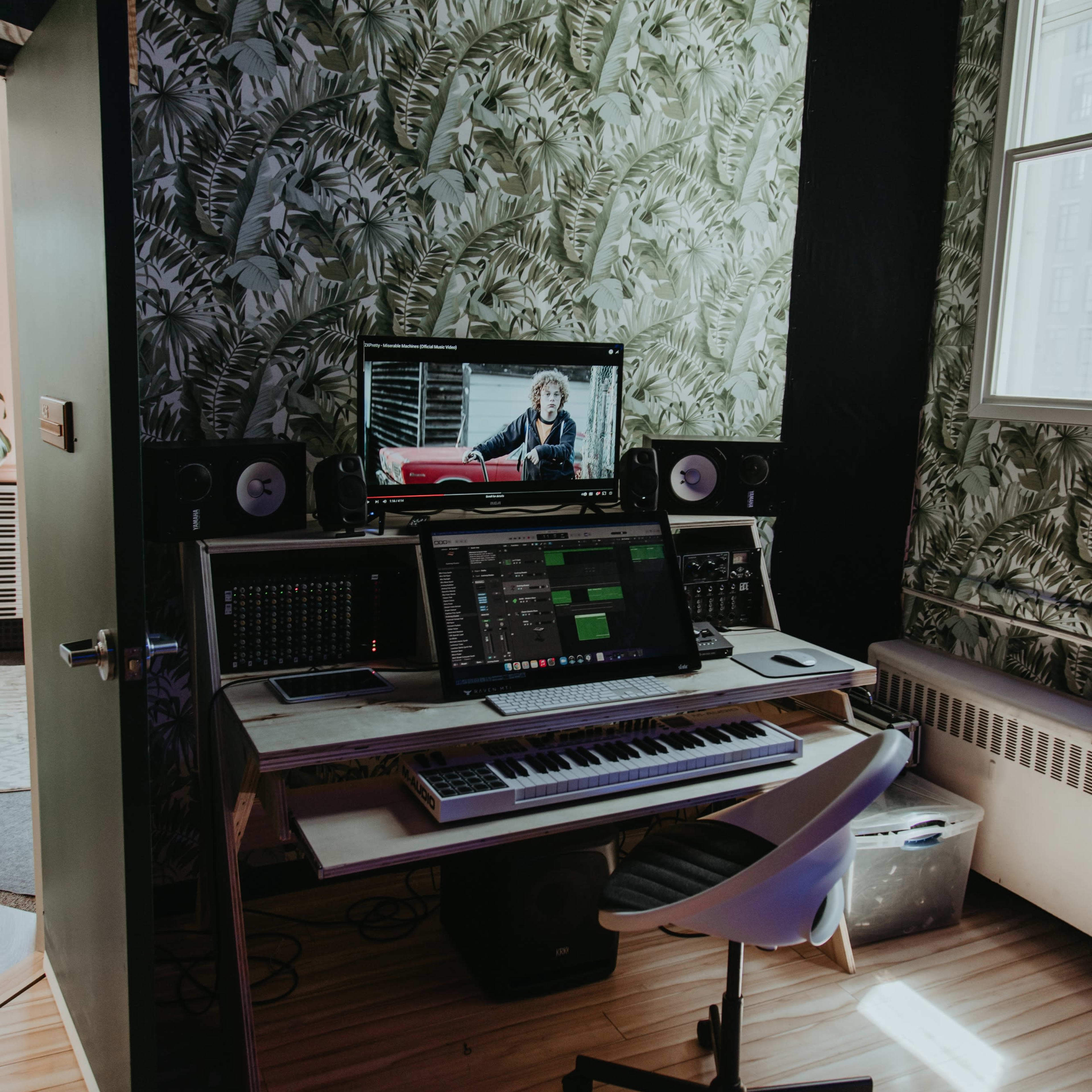 A music production workspace features a computer monitor with a video, a keyboard, and audio equipment against a backdrop of leafy wallpaper.