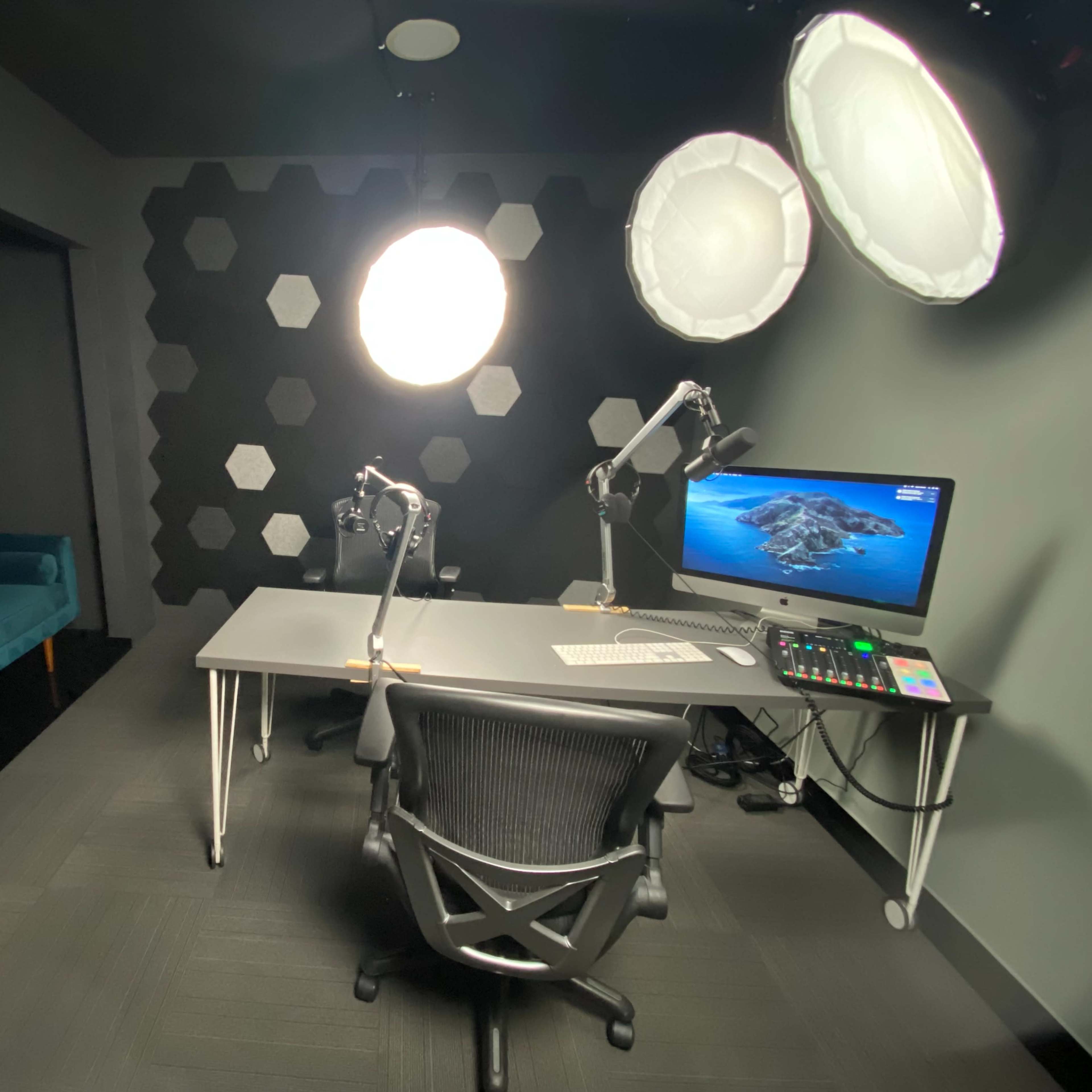 A modern studio setup with a desk, two microphones, a computer monitor displaying an island scene, and a teal sofa against a textured wall.