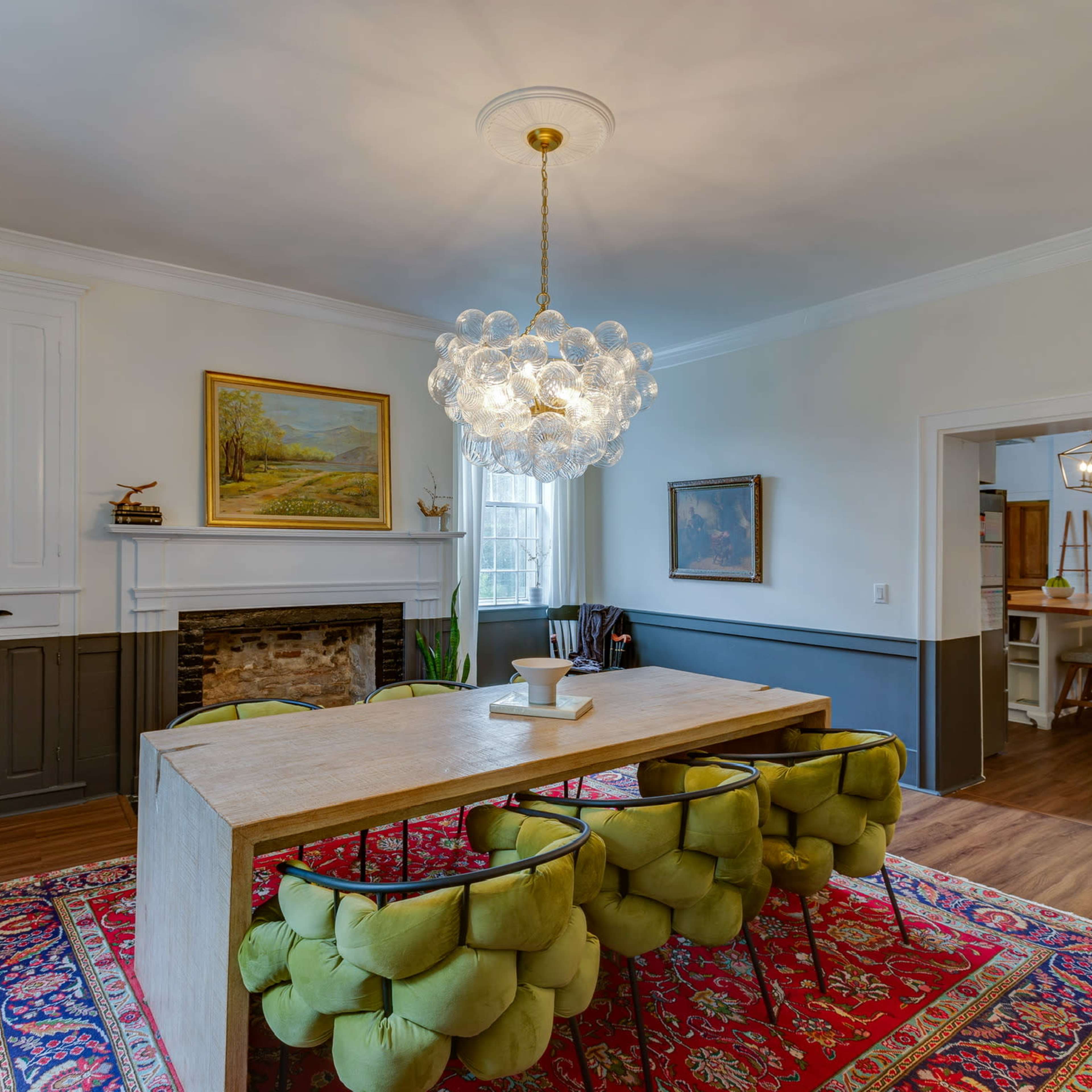 A dining room features a large rectangular table surrounded by green upholstered chairs, a chandelier overhead, and artwork and a fireplace along the walls.