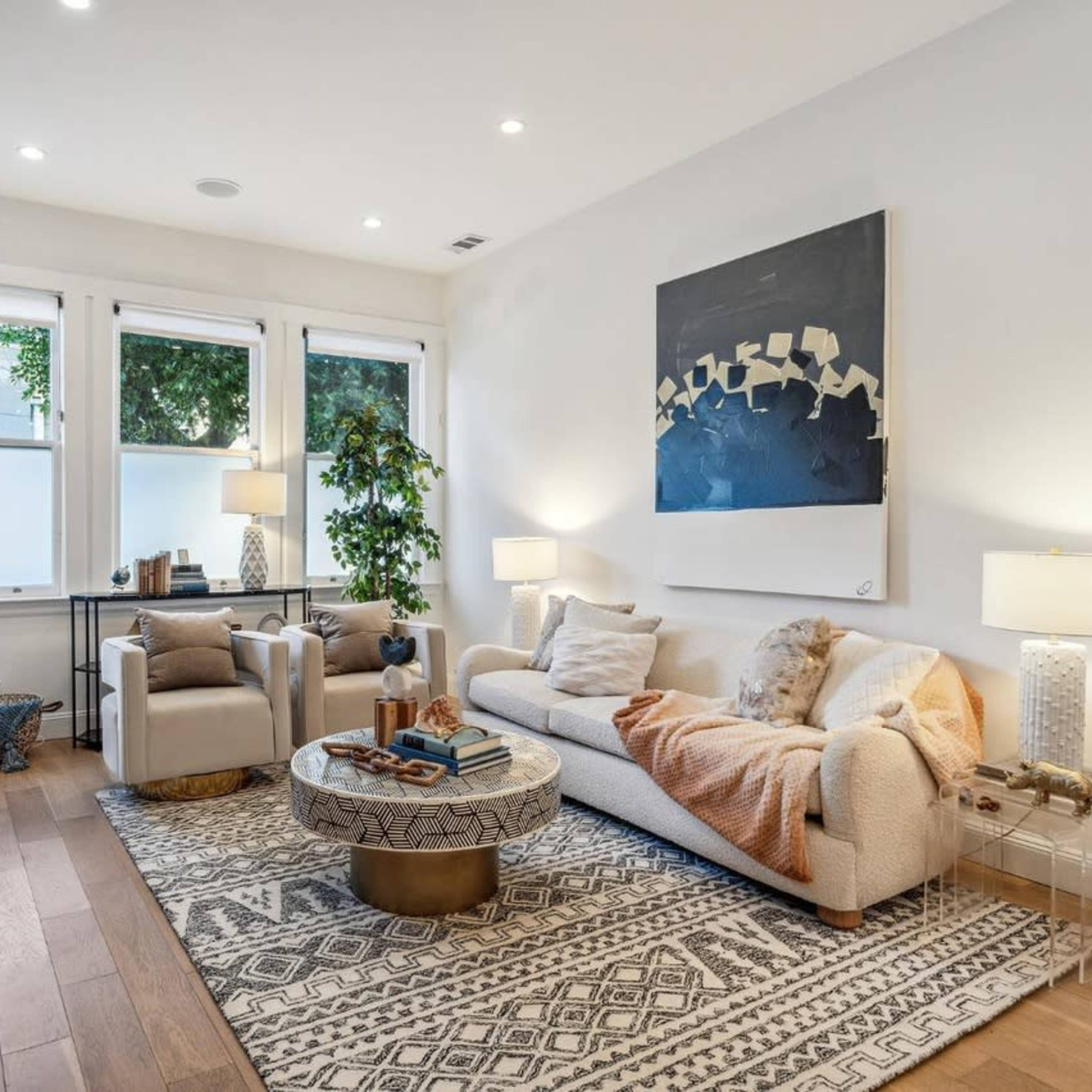 A well-lit living room featuring a beige sofa, two armchairs, a round coffee table, and large windows with greenery outside.