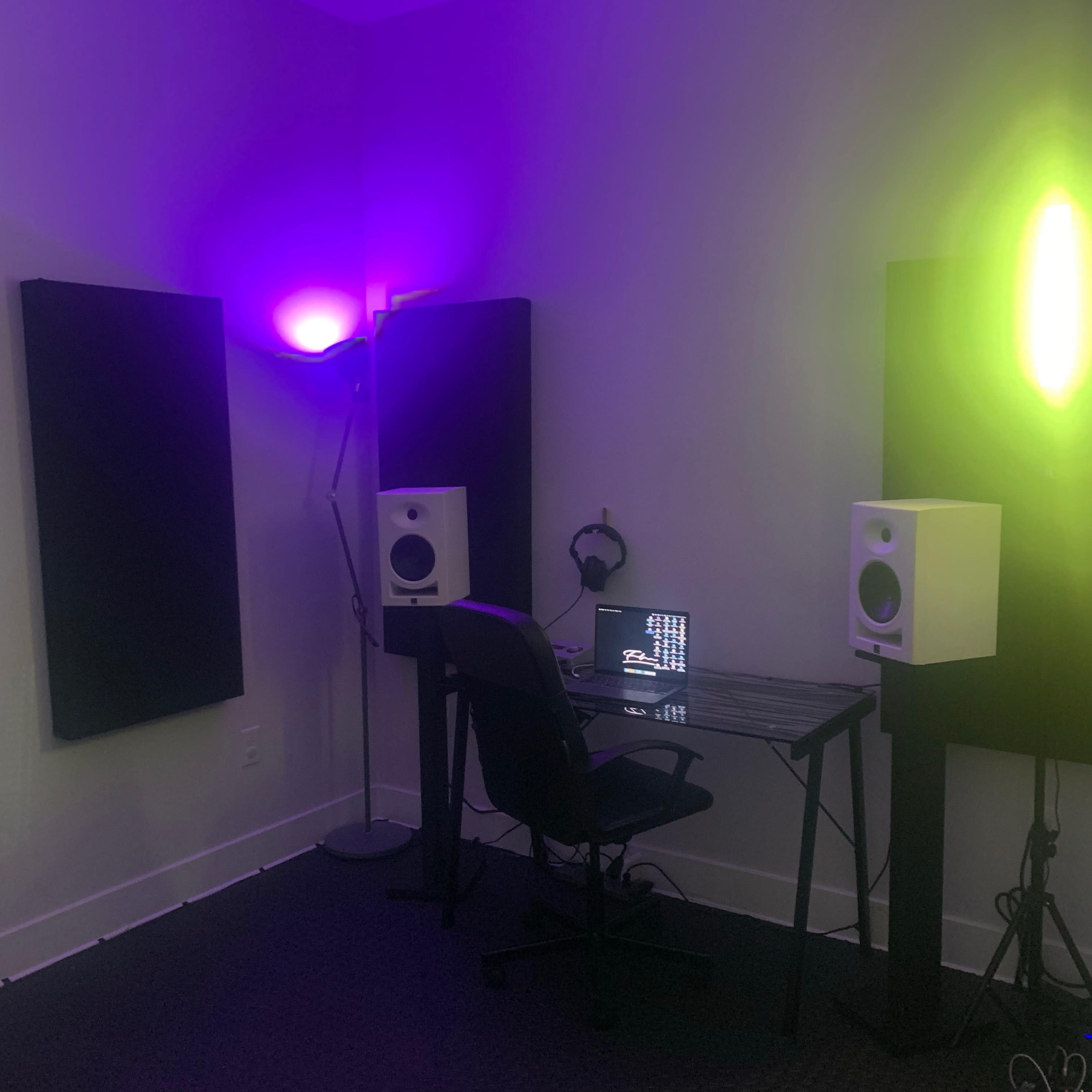 A small music studio is set up with two speakers, a computer on a desk, and acoustic panels on the walls, illuminated by colored lights.