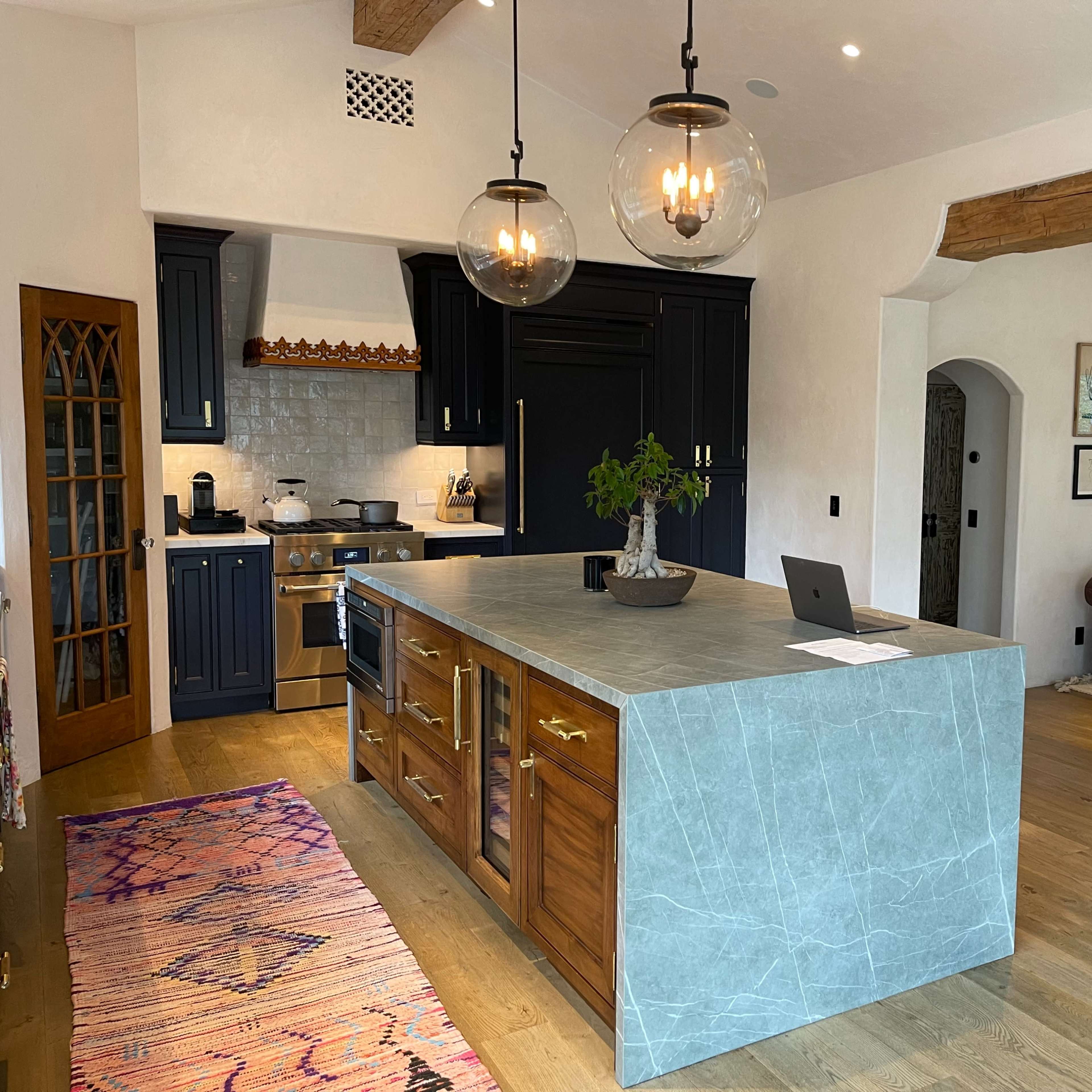 A modern kitchen with a large island featuring a mix of wooden and stone materials, surrounded by a living area with a brown leather sofa and framed pictures on the walls.