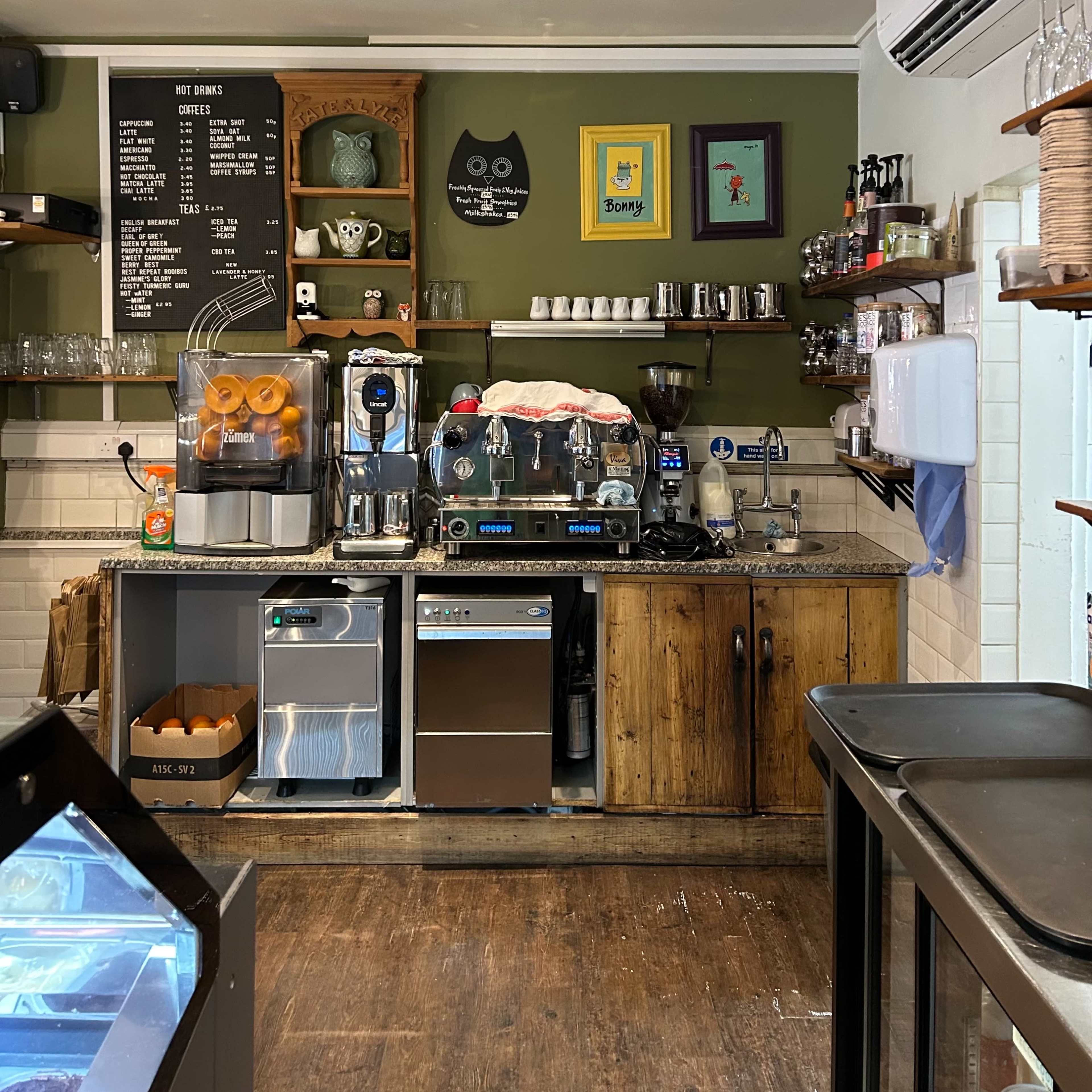 A coffee shop counter featuring an espresso machine, a display of oranges, shelves with glassware, and decorative wall art.