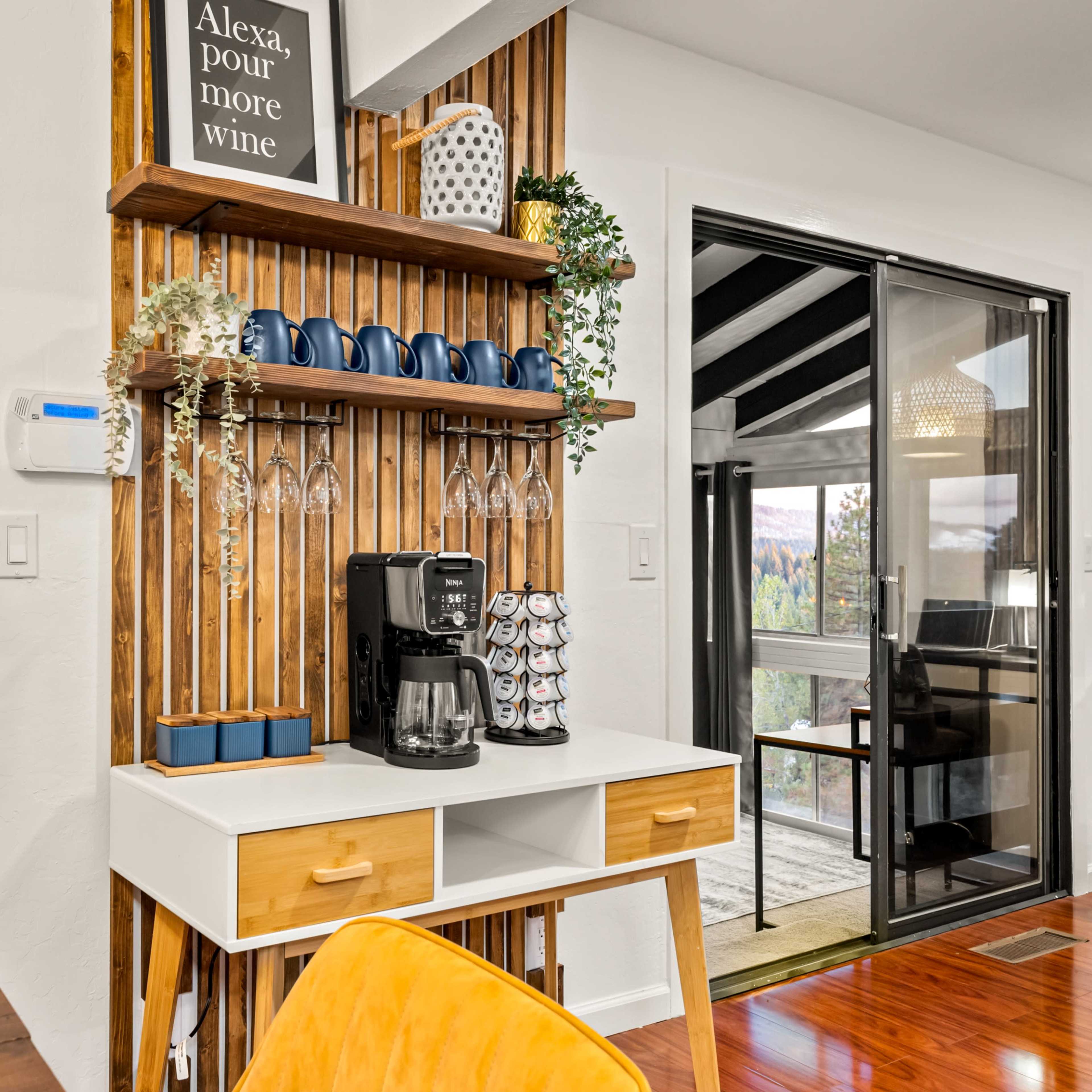 A modern kitchen corner features a coffee maker, a storage shelf with glassware, and a decorative sign, with large windows revealing a forest view.