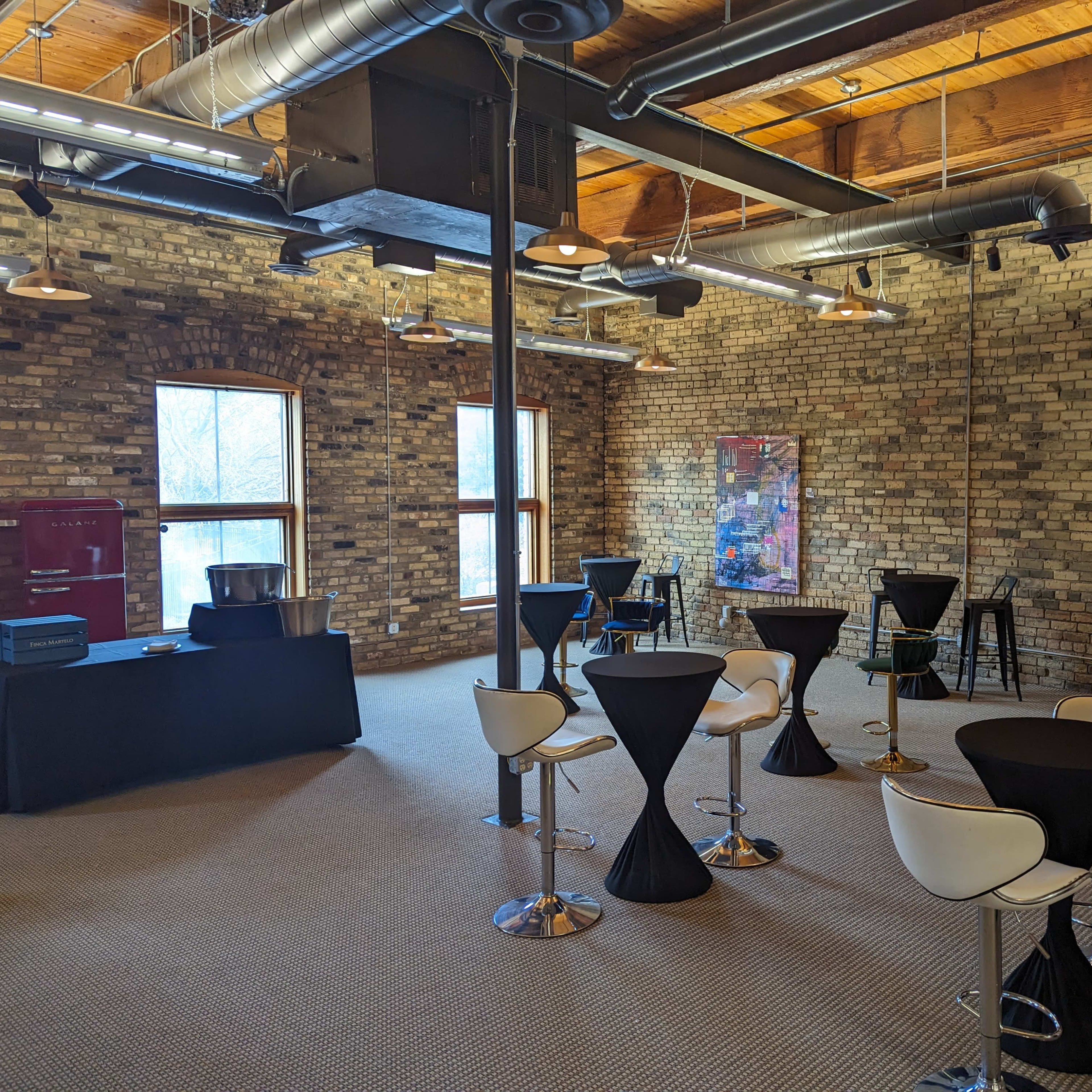 A spacious indoor event space with exposed brick walls, several high-top tables, and a service station in one corner.