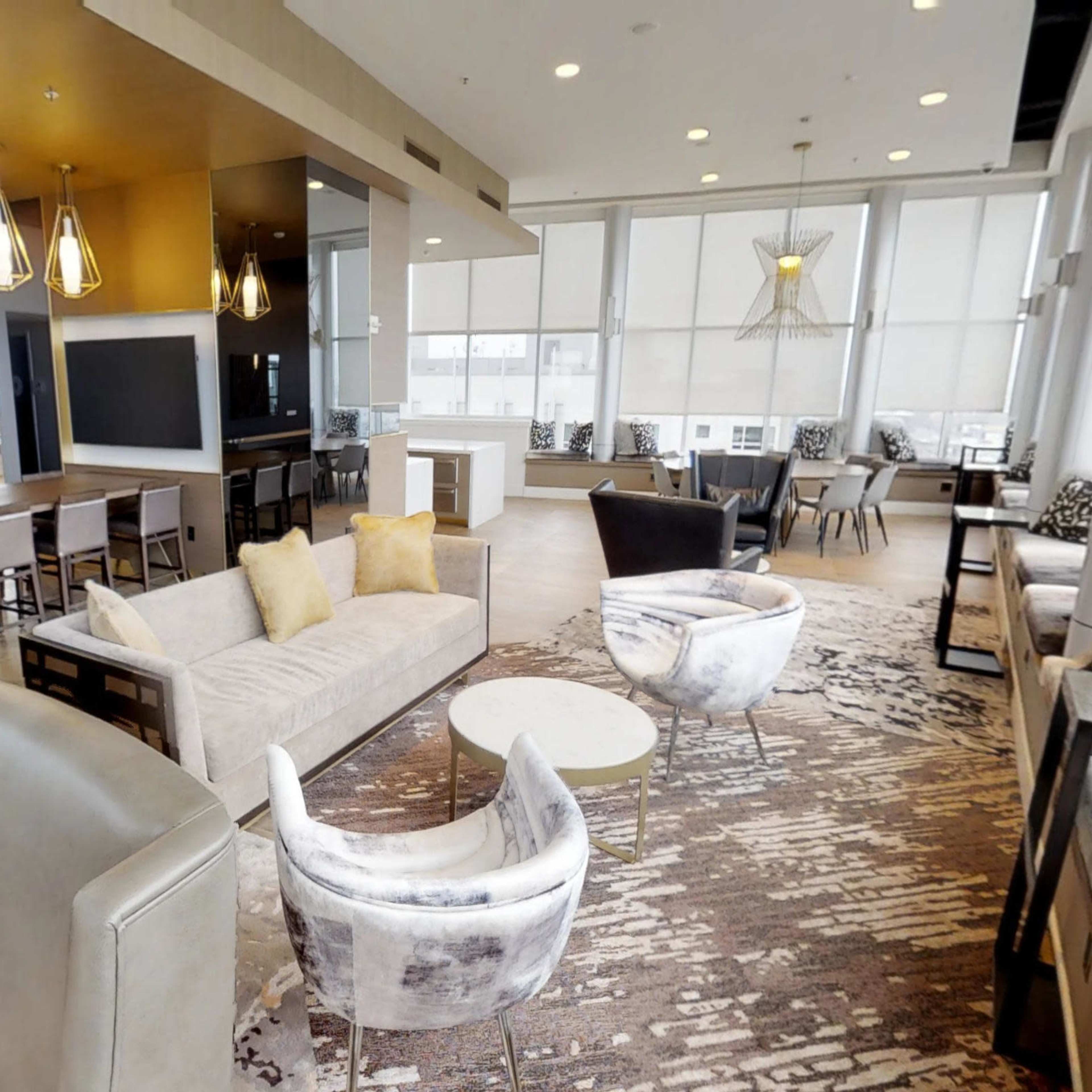 A modern lounge area features comfortable seating, large windows with city views, and a dining table.